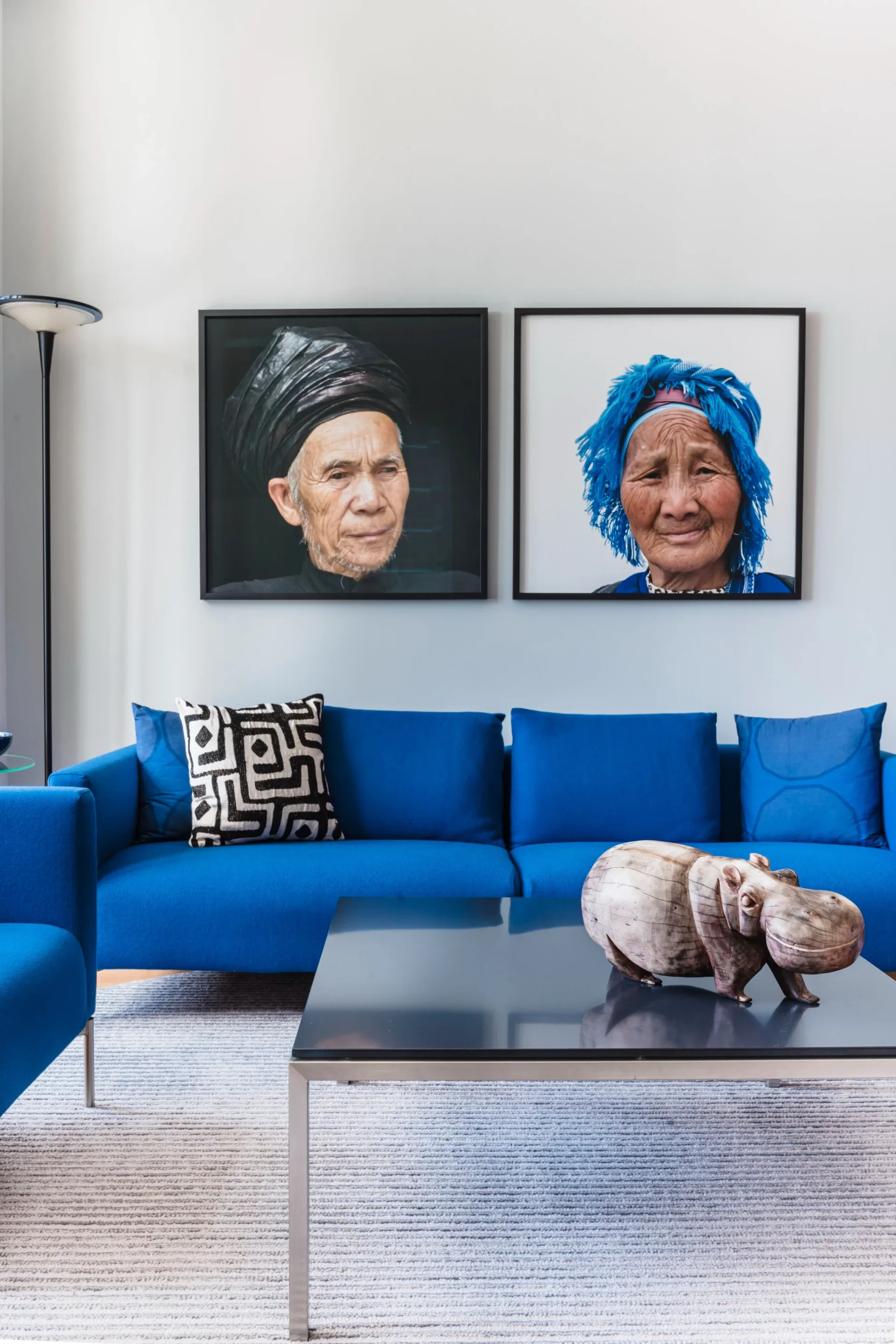 Modern living room with vibrant blue sectional sofa, framed portrait artworks above, geometric patterned pillow, and sculptural hippopotamus accent on glass coffee table by Alina Hackett.