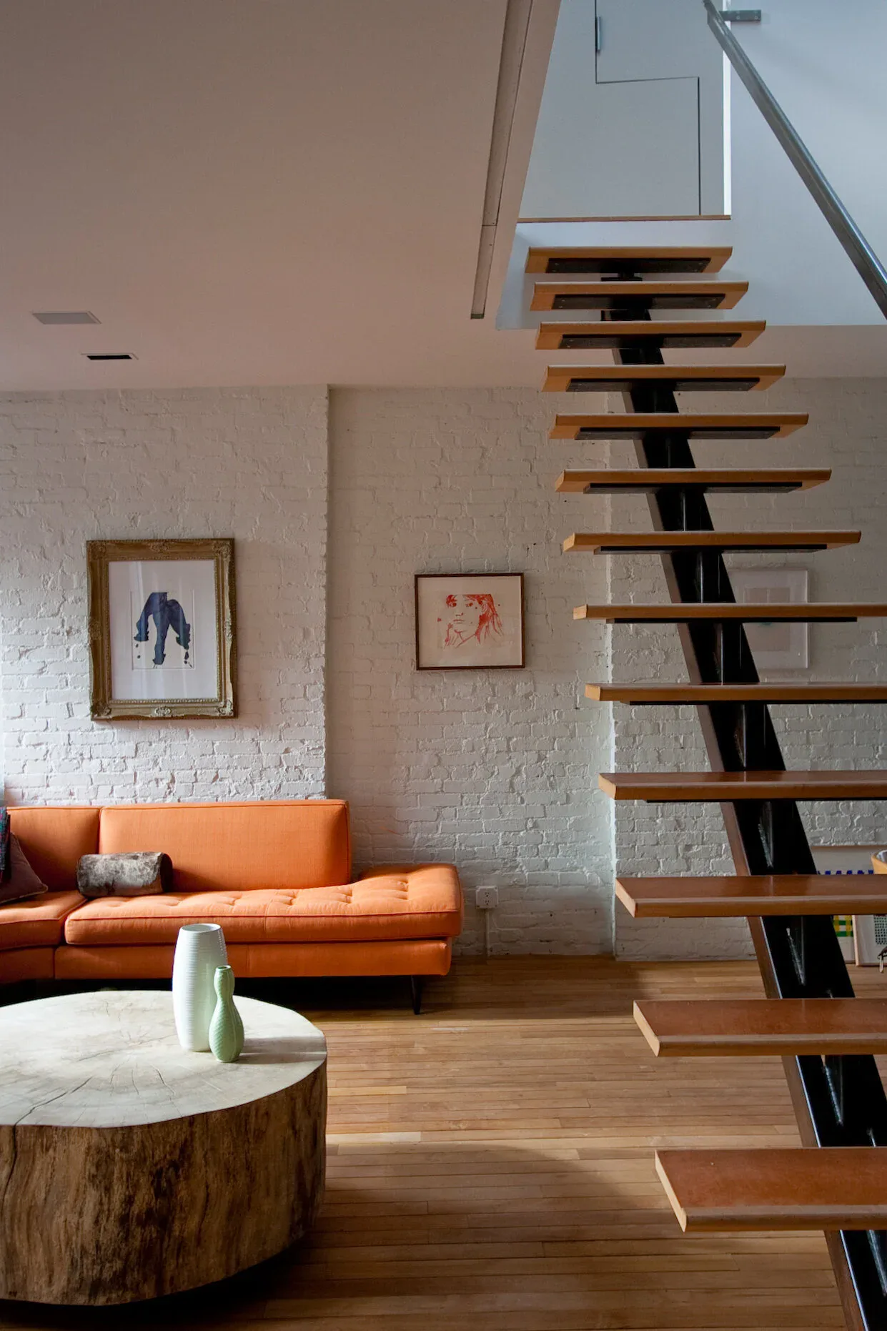 Modern loft interior featuring white brick walls, orange mid-century sofa, wooden round side table, and floating wooden staircase with black steel frame by Alina Hackett.