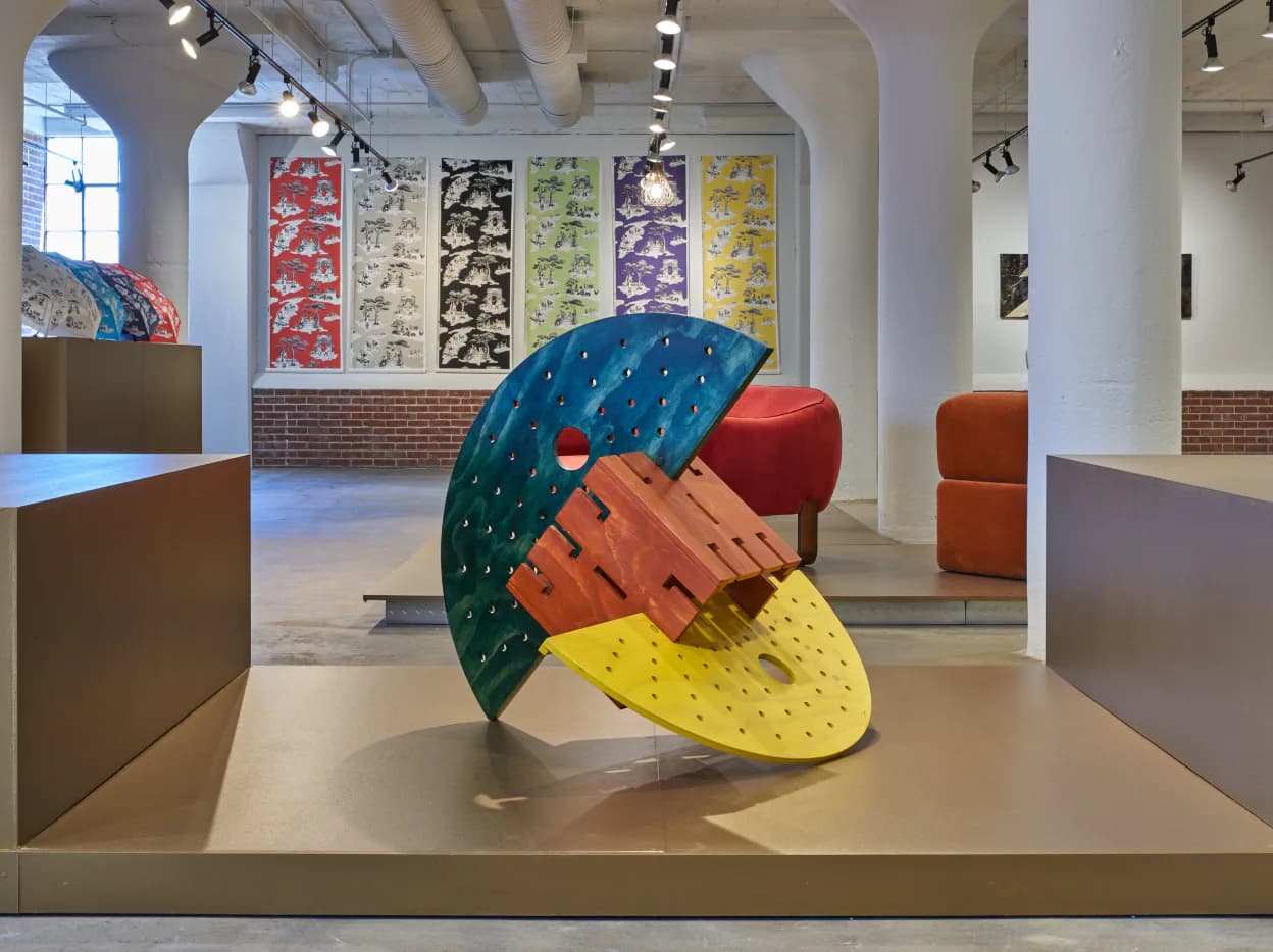 Gallery interior with colorful geometric sculptural installation featuring curved forms in teal, red, orange, and yellow, with patterned textile panels on walls, by Alina Hackett.