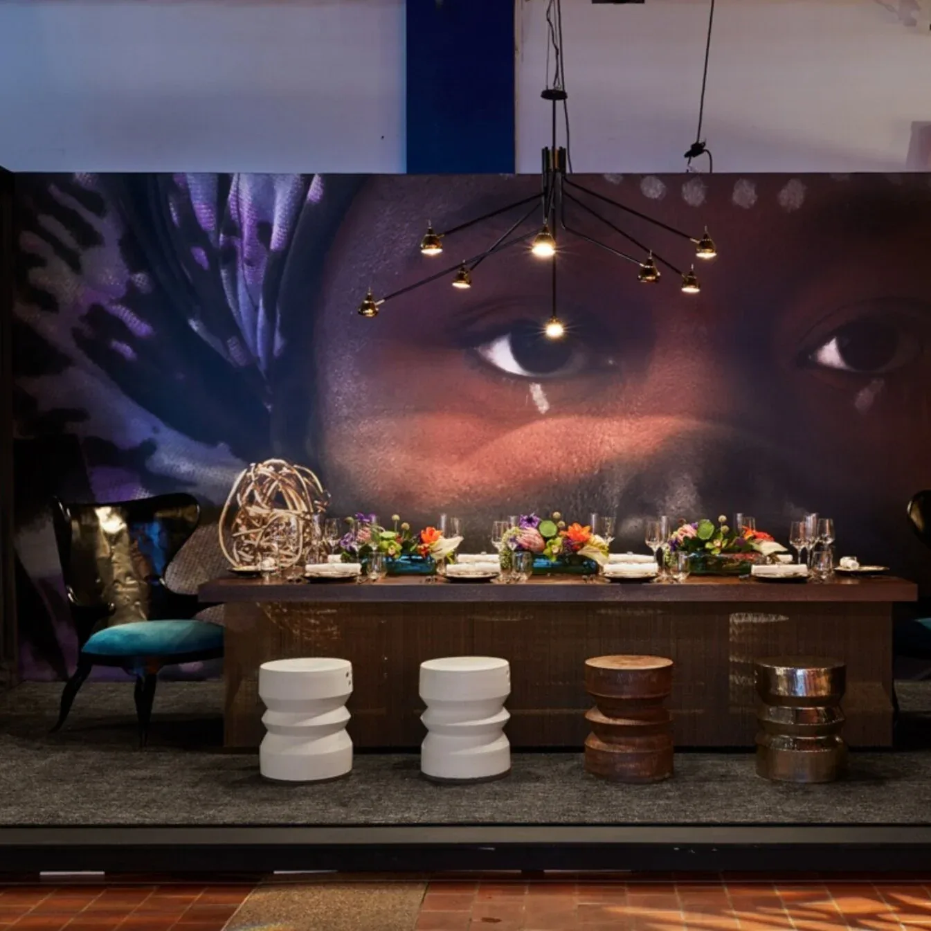 Modern dining installation with dark wood table, white sculptural stools, hanging pendant lights, and dramatic backlit mural, by Alina Hackett.