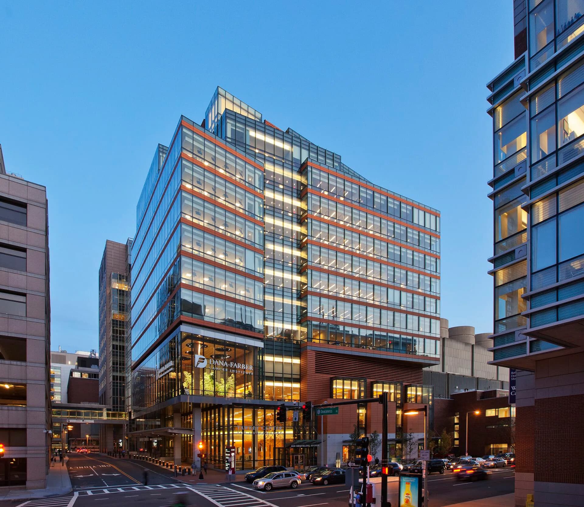 Contemporary mixed-use building with brick and glass facade, illuminated ground-floor retail spaces, and residential towers above at dusk by Alison Fidler.
