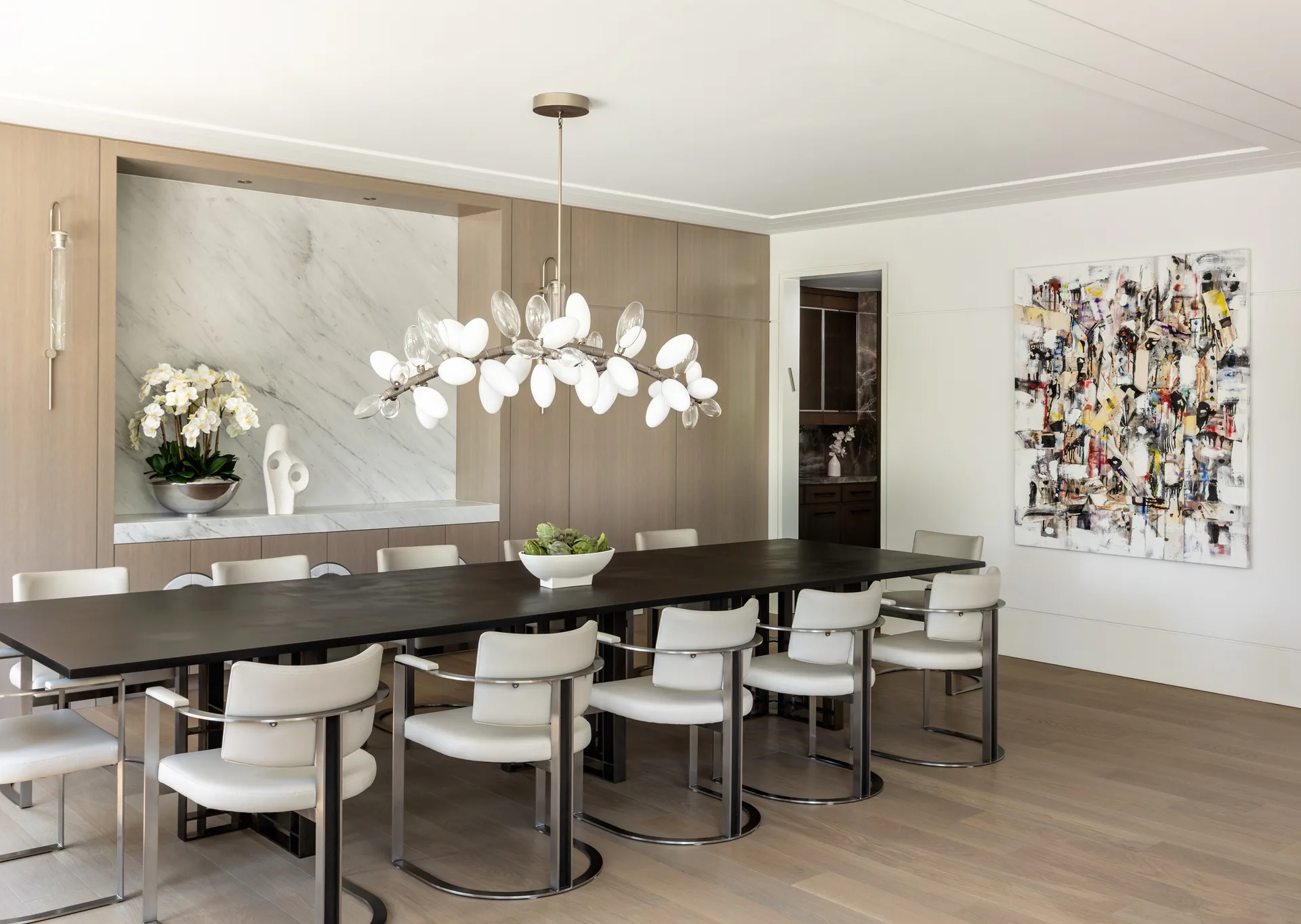 Modern dining room with black table, cream curved chairs, and sculptural pendant light, neutral palette with marble accent wall by Aliza Katz.