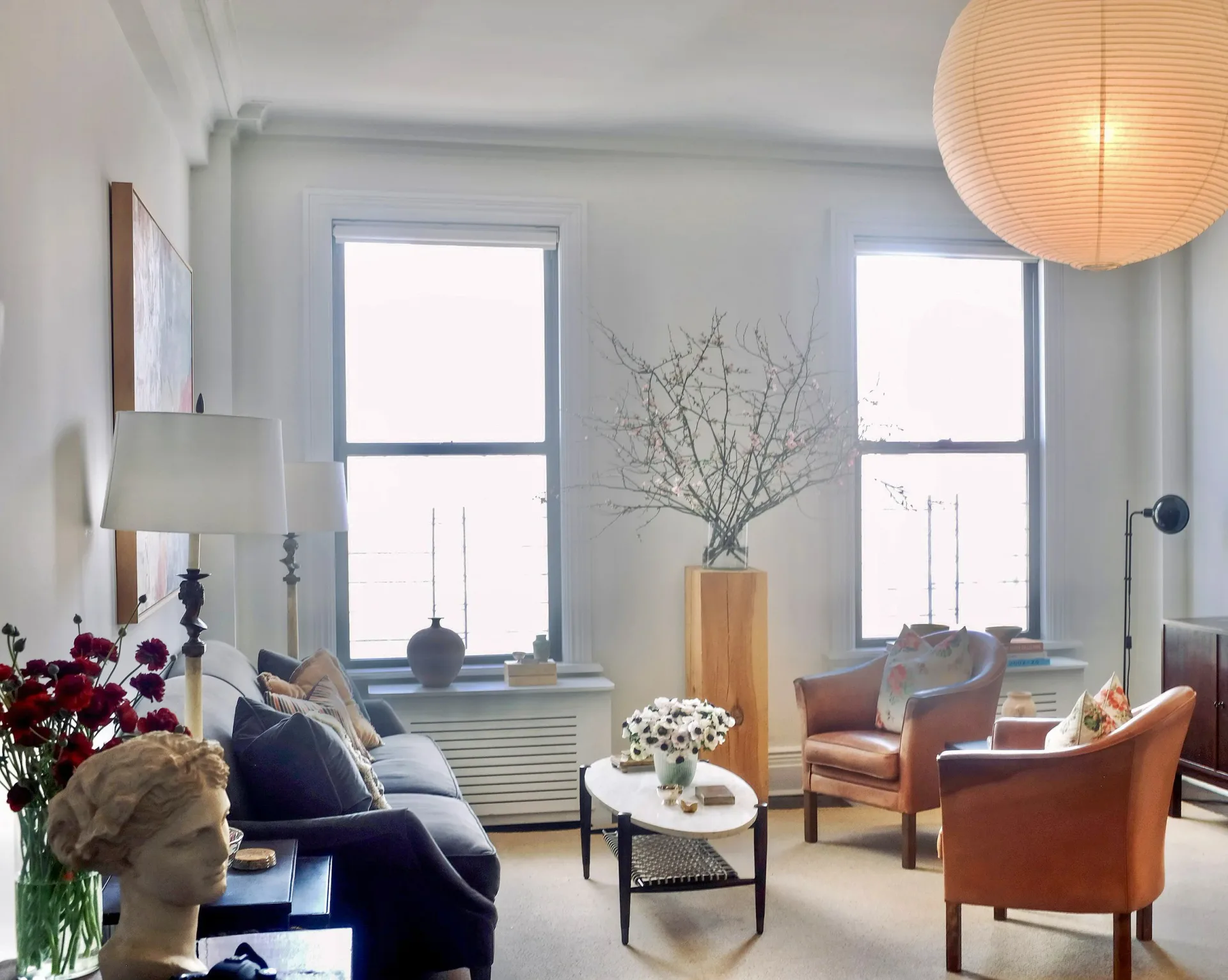 Bright living room with two large windows, terracotta armchairs, navy sofa, wooden vase with branches, and pendant lighting, designed by Allison Alexander.