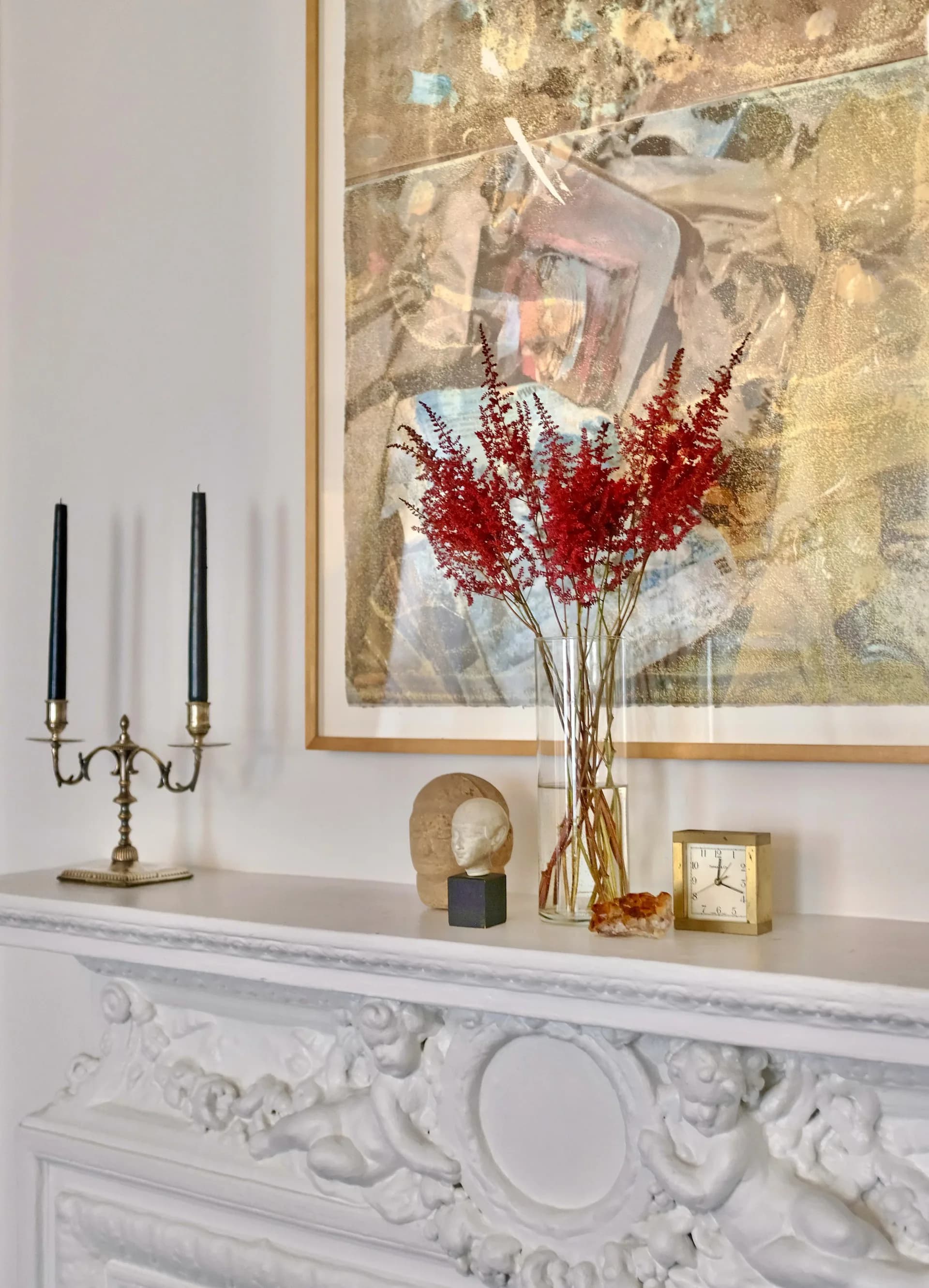Mantelpiece styling with ornate white fireplace, brass candelabra, red dried flowers in glass vase, and abstract framed artwork by Allison Alexander.