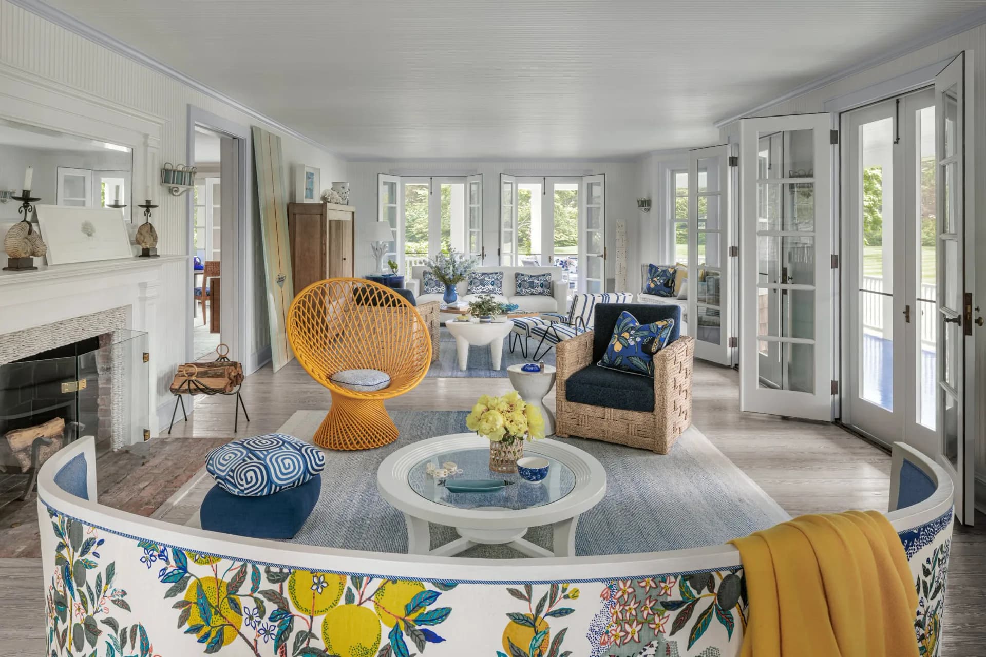 Bright coastal living room featuring a curved blue and yellow patterned sofa, woven rattan chair, and round marble table, with French doors opening to ocean views, designed by Allison Babcock.