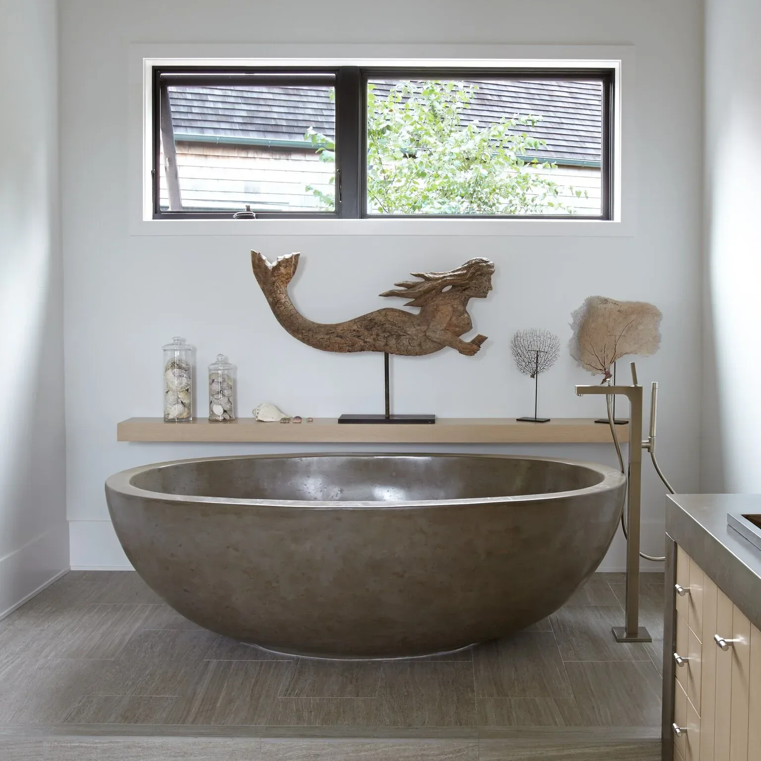 Modern bathroom with sculptural bronze soaking tub, floating vanity, decorative wooden mermaid sculpture, and natural light from transom windows by Allison Babcock.
