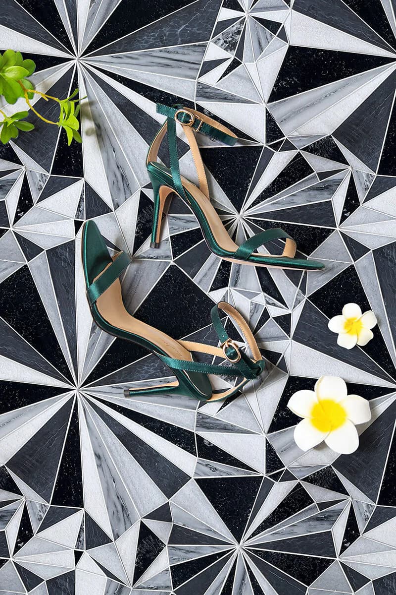 Pair of emerald green heels with tan accents displayed on a geometric black and white patterned surface, styled with white flowers and greenery by Allison Eden.