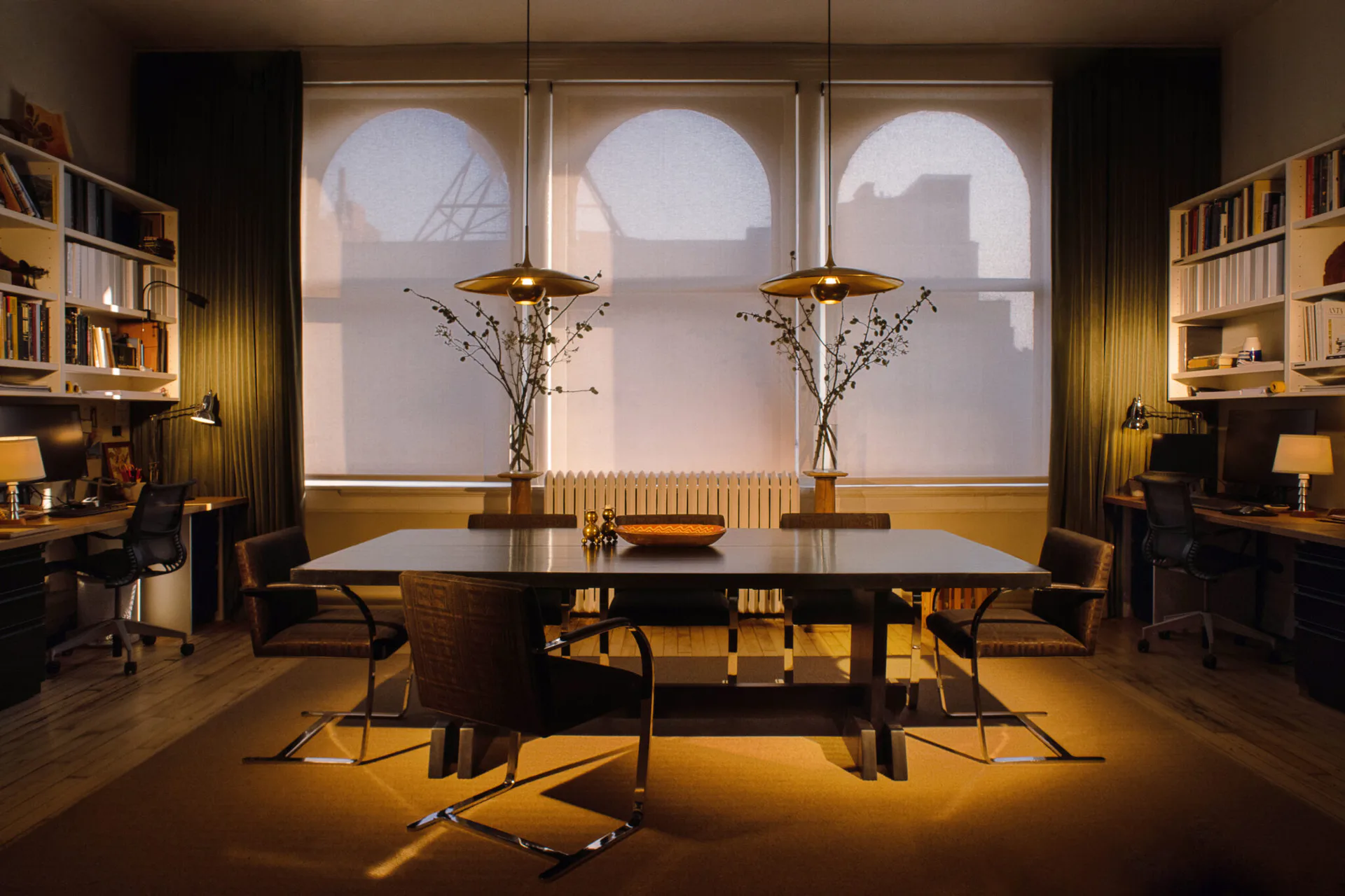 Sophisticated dining room with dark table, brass pendant lights, arched windows, warm golden lighting, bookshelves, and neutral palette by Alyssa Nguyen.