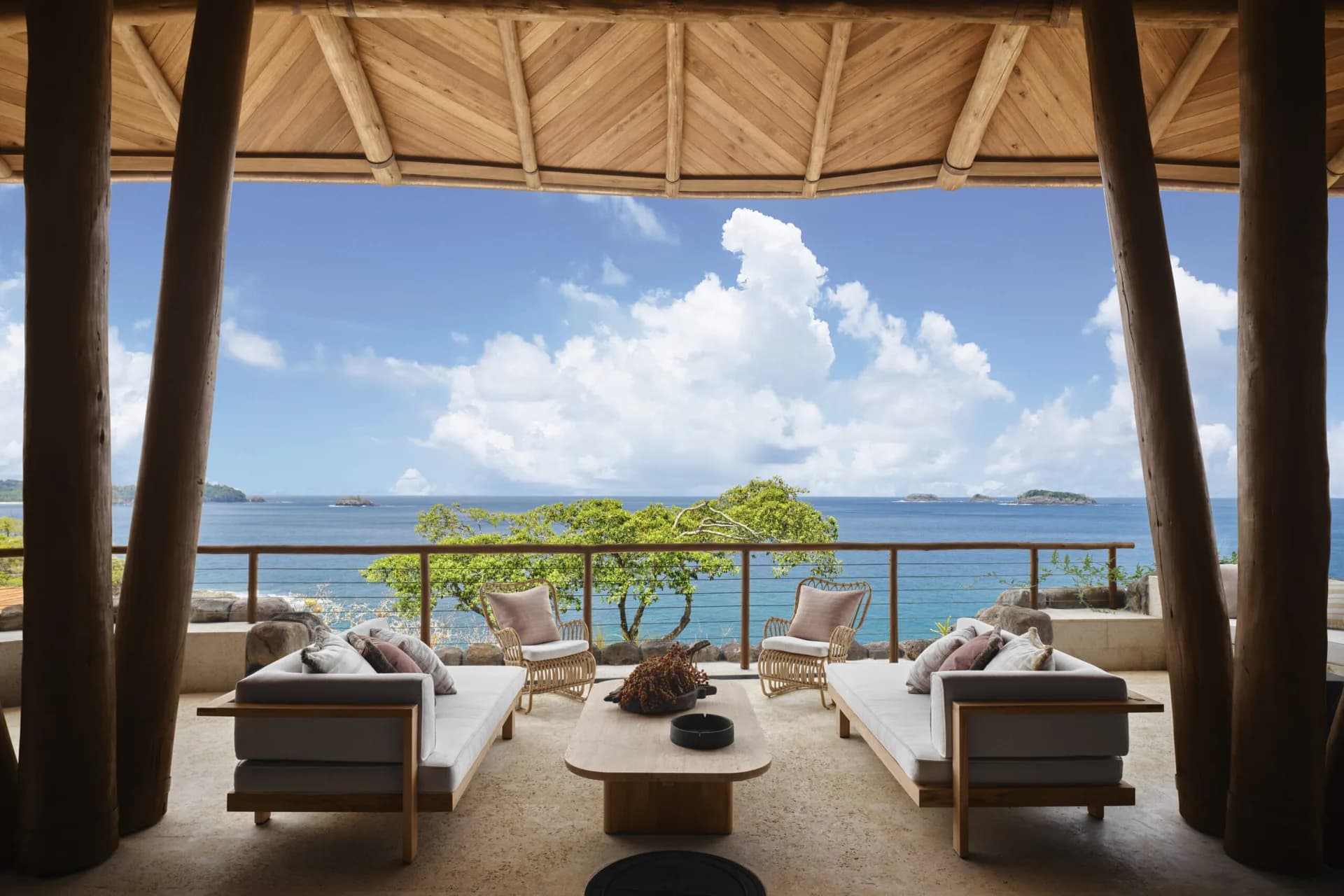 Luxurious open-air pavilion with wooden structural beams overlooking a turquoise ocean, featuring neutral furnishings and a serene tropical setting by Alyssa Nguyen.