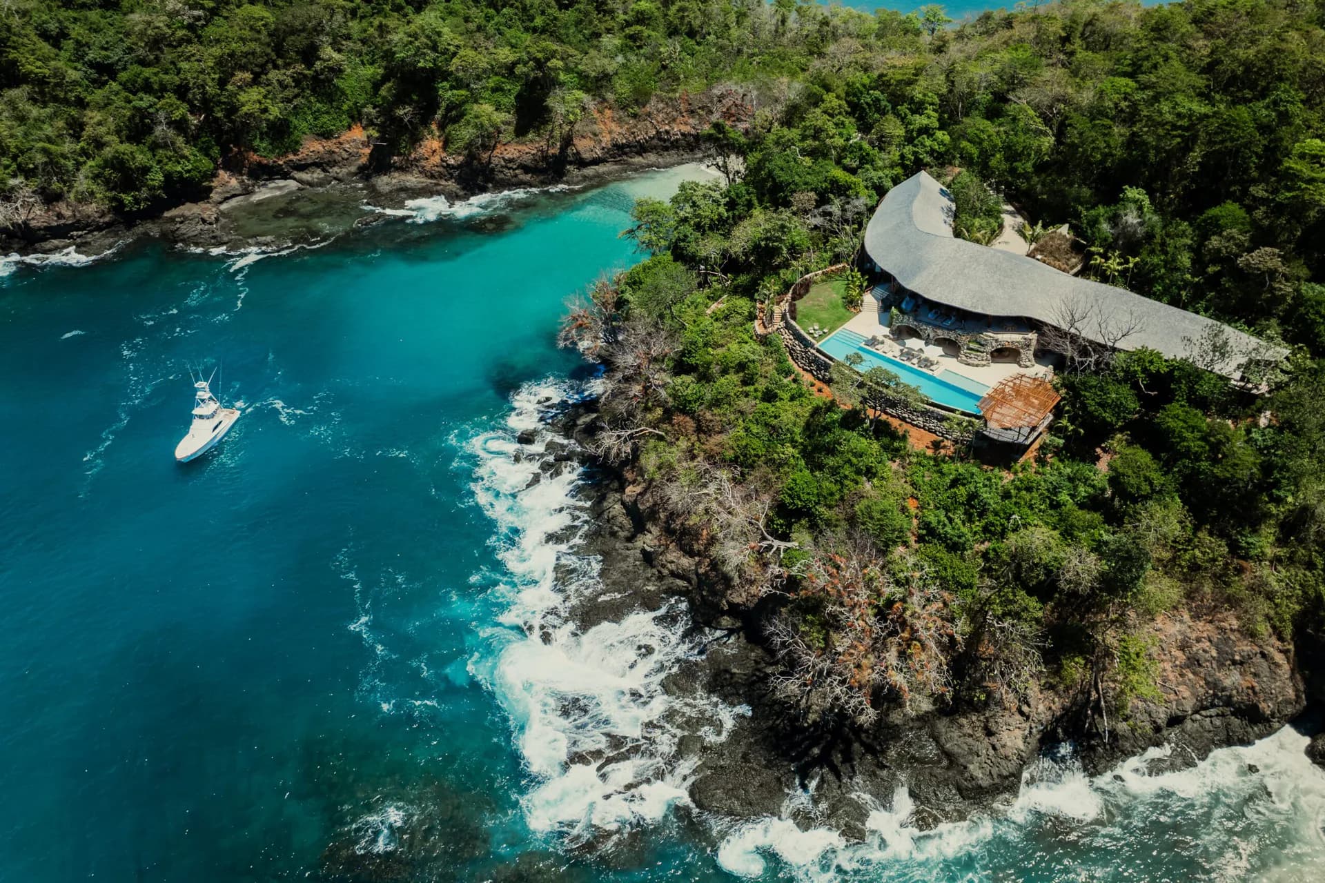 Curved modernist structure perched on a dramatic rocky coastal cliff overlooking turquoise ocean waters surrounded by lush tropical vegetation, designed by Alyssa Nguyen.