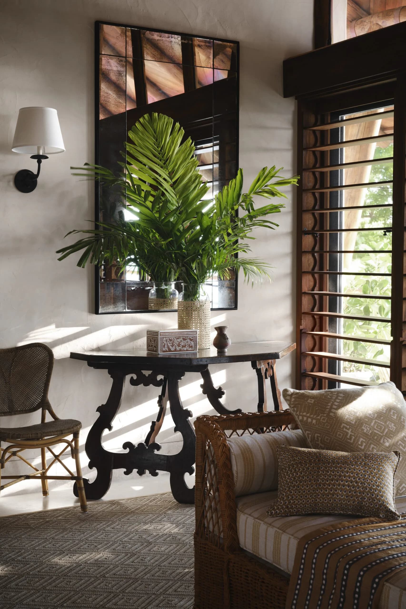 Bright residential interior with black ornamental table, potted palm plant, woven rattan chair, striped upholstered seating, and natural light from louvered doors by Alyssa Nguyen.
