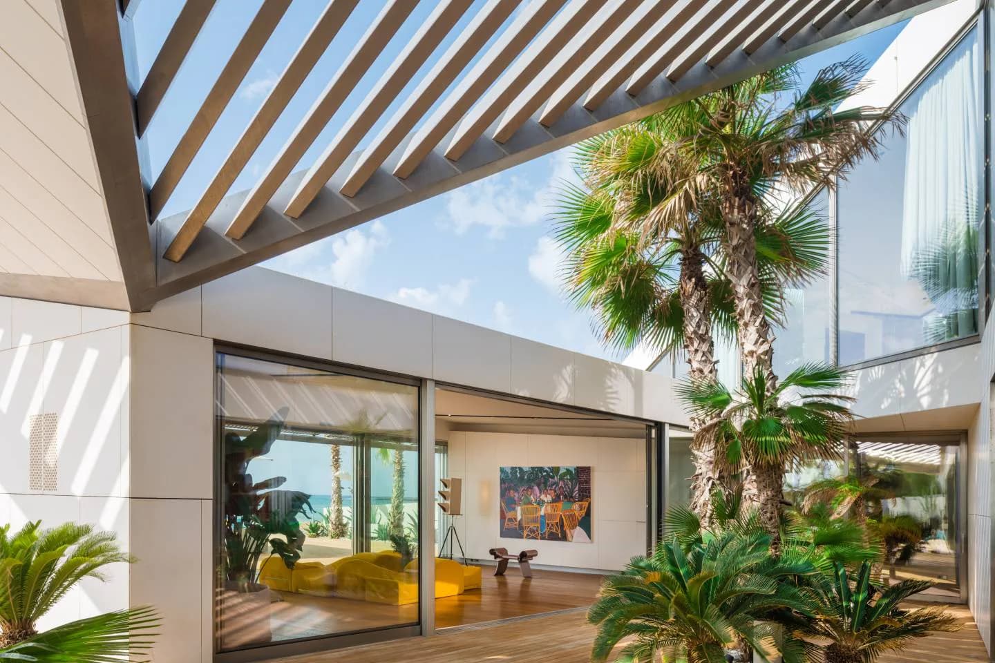 Modern residential courtyard with slatted wooden pergola, floor-to-ceiling glass doors, tropical plants, and wooden deck flooring, designed by Amale Andraos.