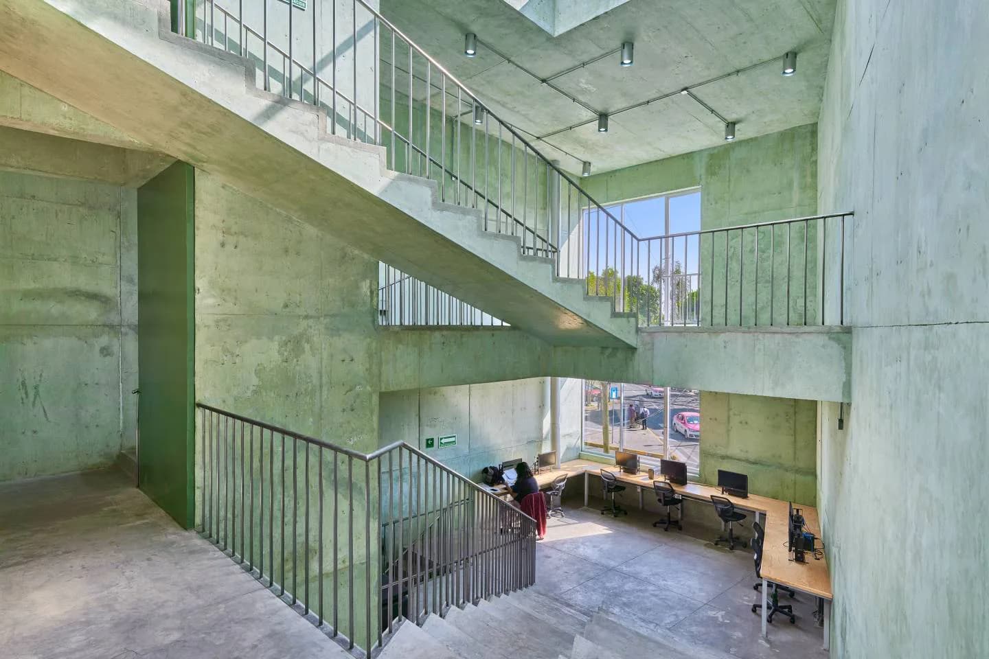 Concrete multi-level interior with metal railings and stairs, featuring a minimalist workspace below, designed by Amale Andraos.