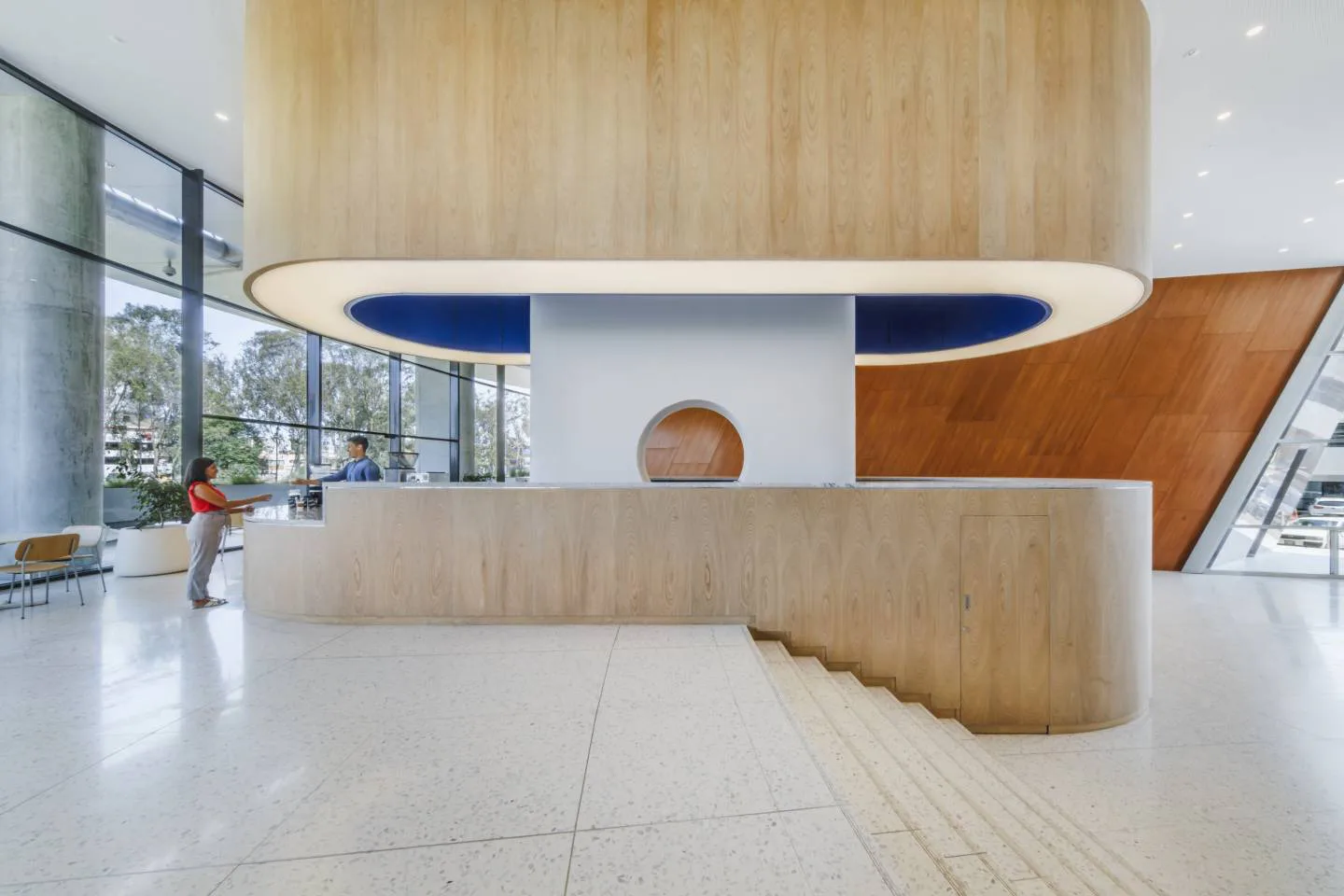 Modern reception area with curved wooden counter, blue and white signage, light wood paneling, and polished concrete floors, designed by Amale Andraos.