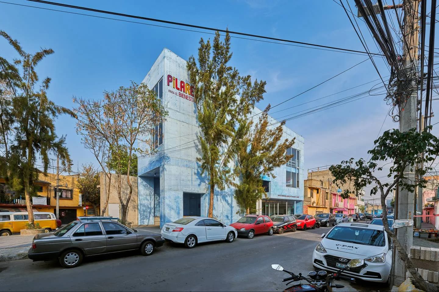 Bright white contemporary building with blue signage on a street corner, surrounded by parked cars and trees, designed by Amale Andraos.