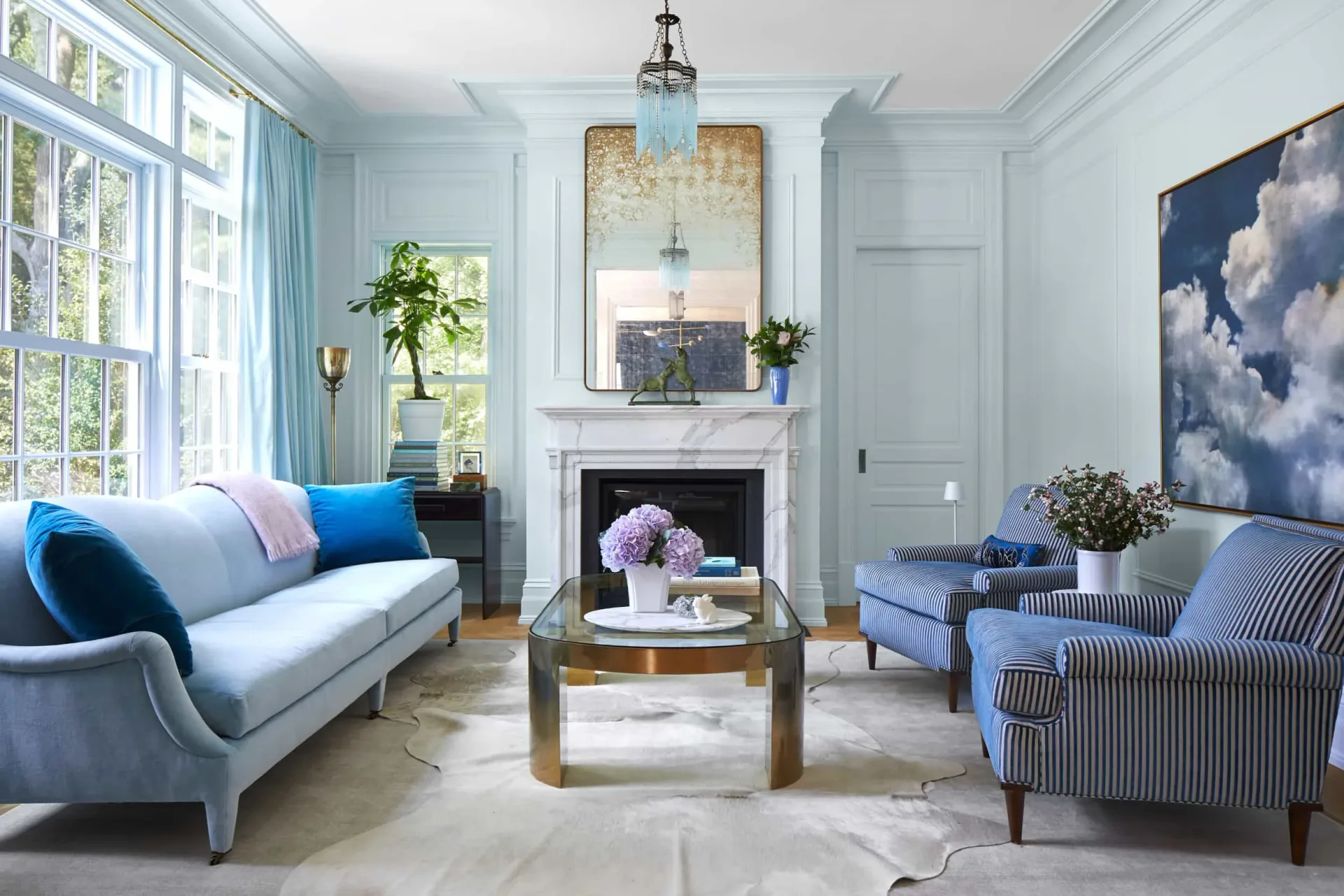 Light-filled living room with pale blue walls, fireplace, and mixed seating in blue and striped fabrics by Amanda Corey.