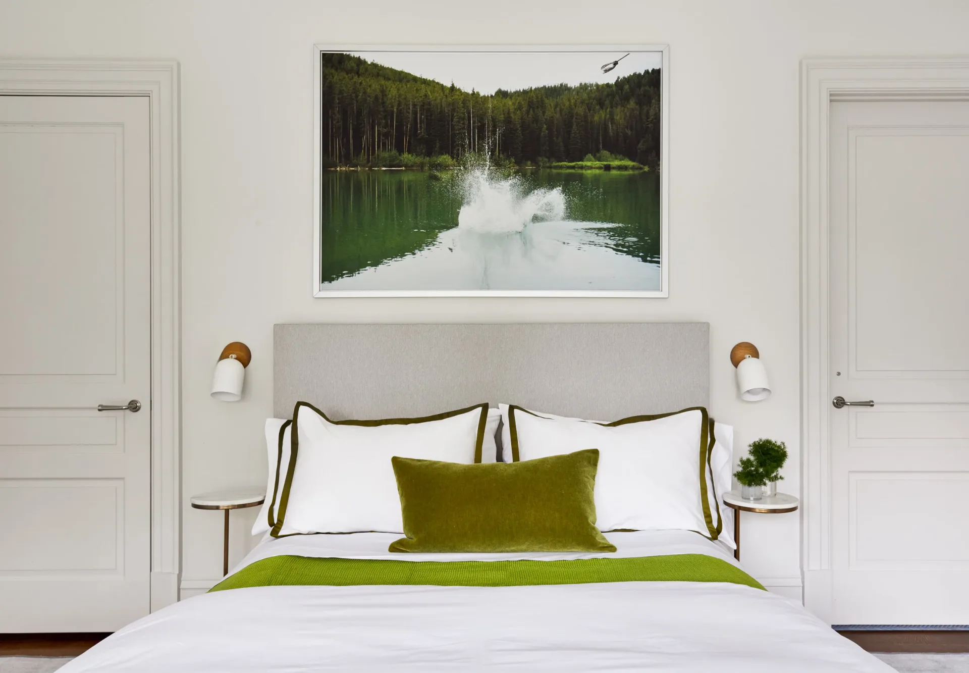Modern bedroom with white upholstered headboard, olive-green accents, brass fixtures, and forest lake landscape artwork above bed by Amanda Corey.