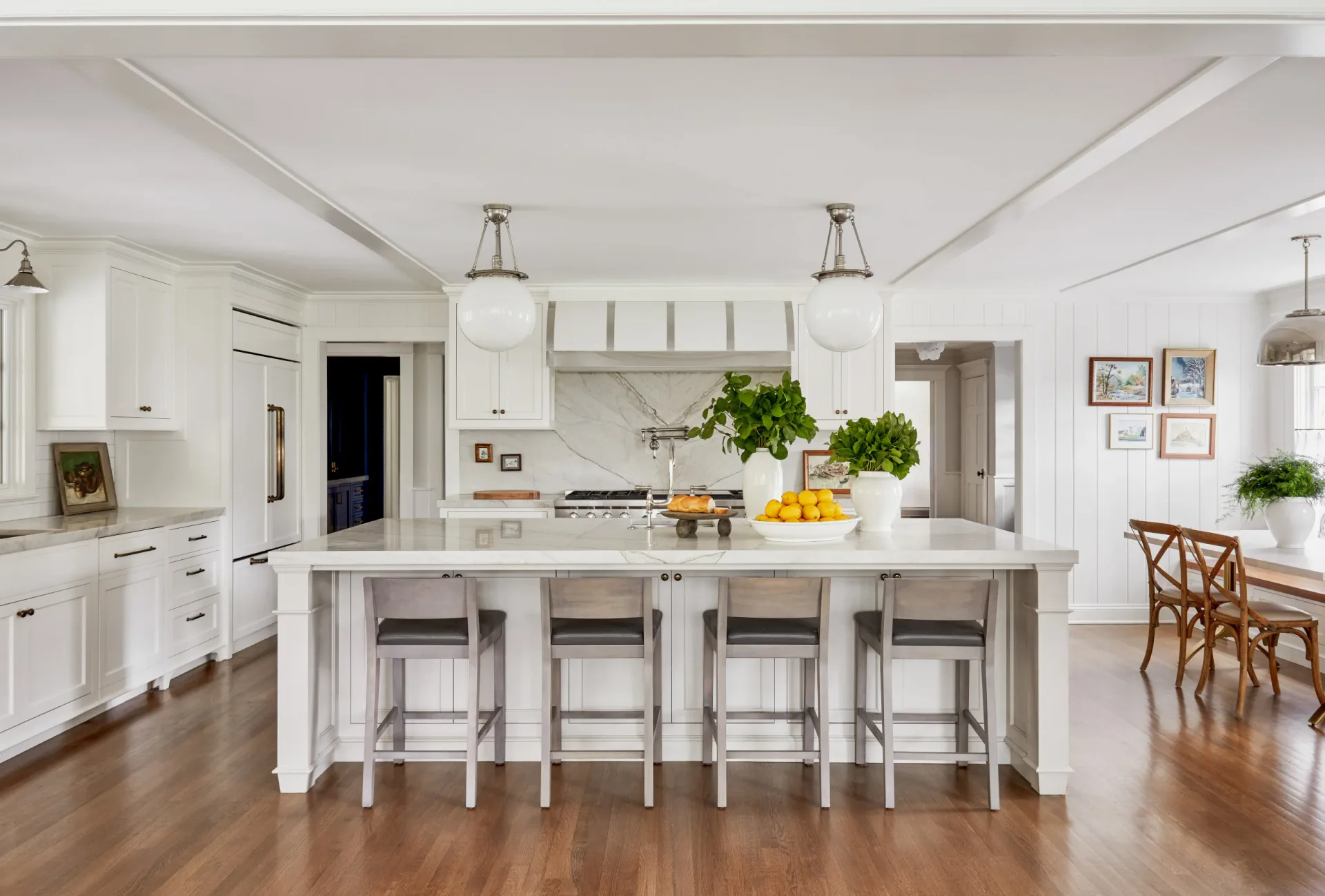 Bright white kitchen with large island, gray upholstered stools, wood floors, and open dining area beyond by Amanda Corey.