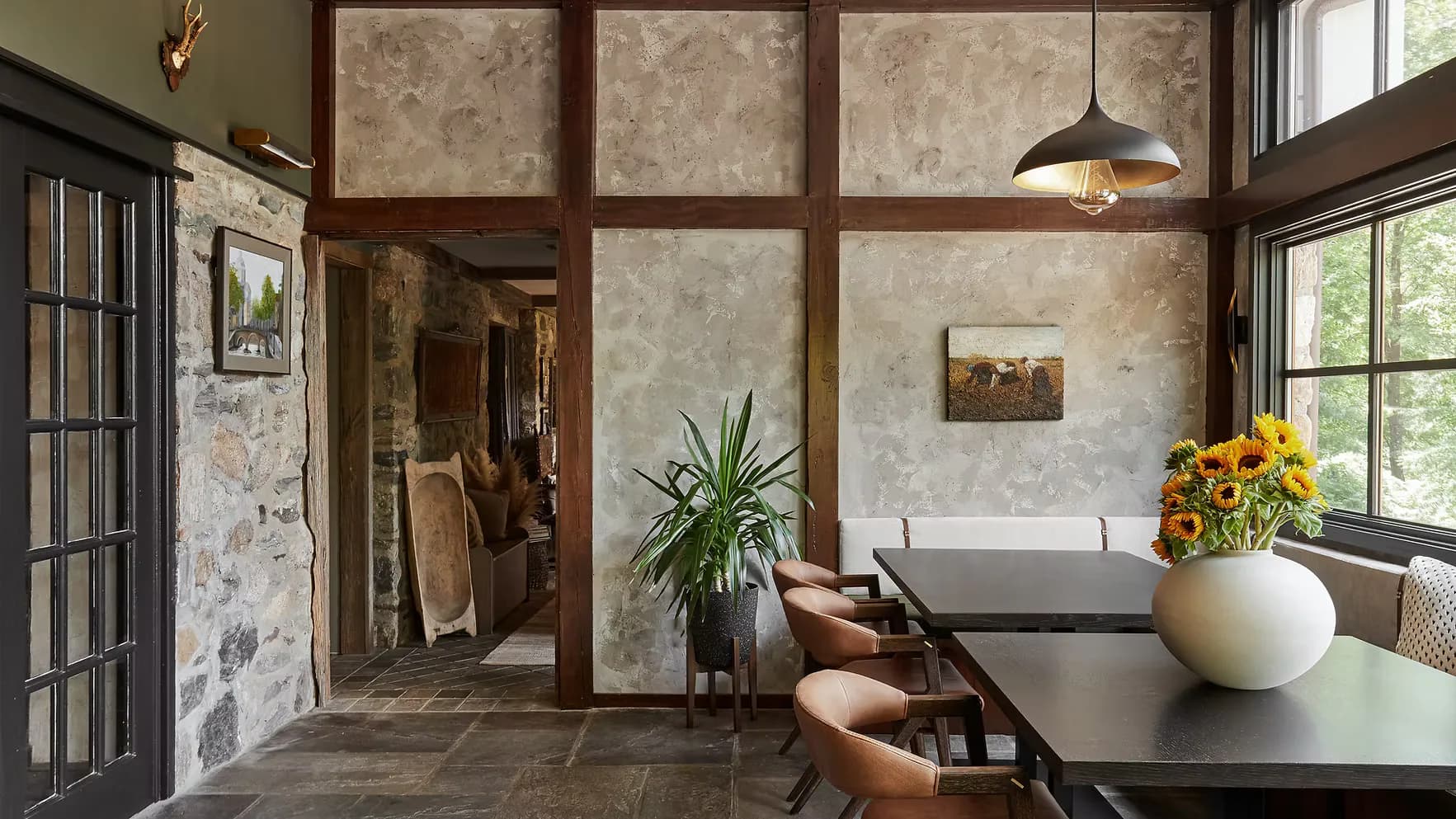 Modern dining space with warm wood-framed glass partitions, concrete flooring, black table, white vessel with sunflowers, and large windows overlooking greenery by Amanda Lehmann.
