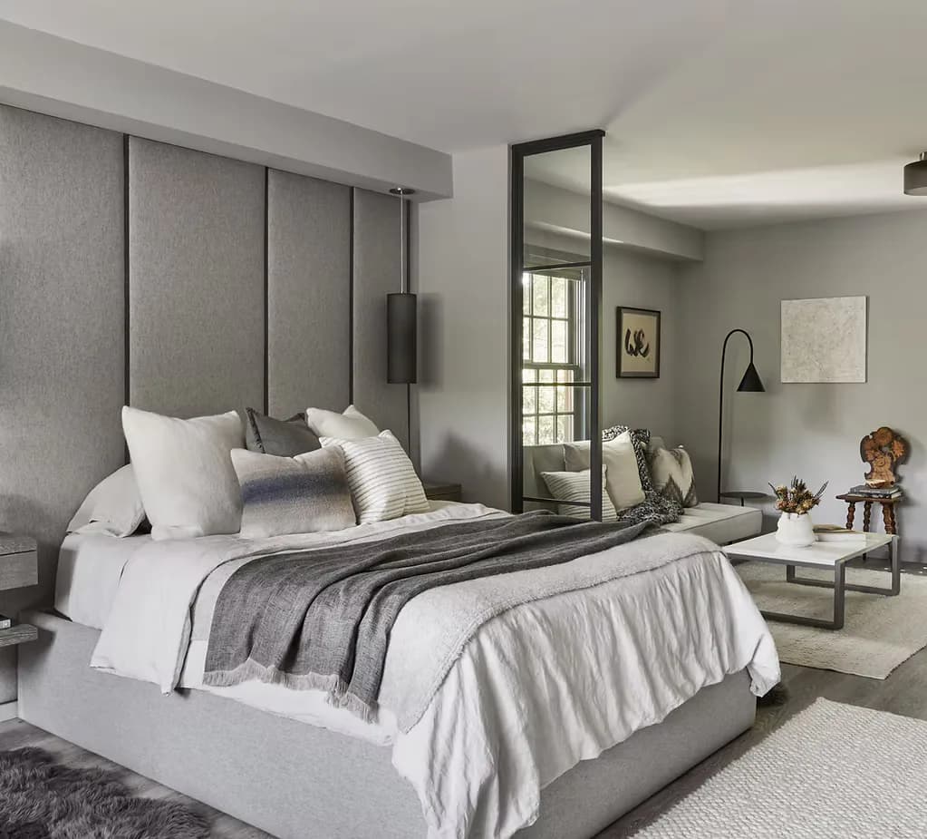 Modern bedroom with gray upholstered headboard, white bedding, layered textiles, full-length mirror, and neutral palette by Amanda Lehmann.