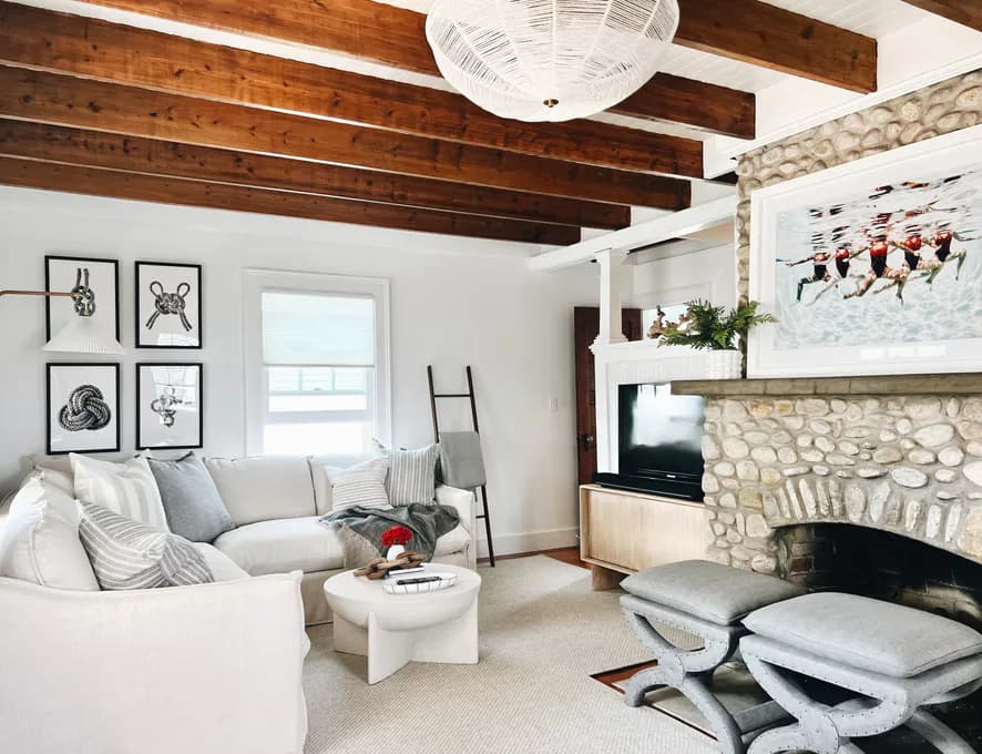 Bright living room with exposed wood beams, stone fireplace, white furnishings, and gallery wall art by Amanda Lehmann.