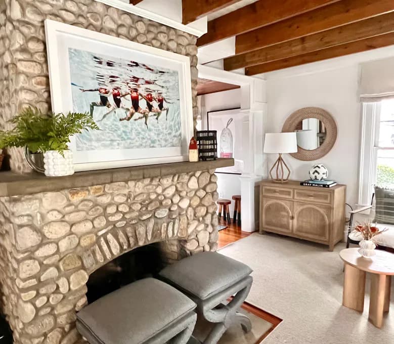 Stone fireplace with framed artwork, wooden beams, and modern furnishings in a light-filled living room by Amanda Lehmann.