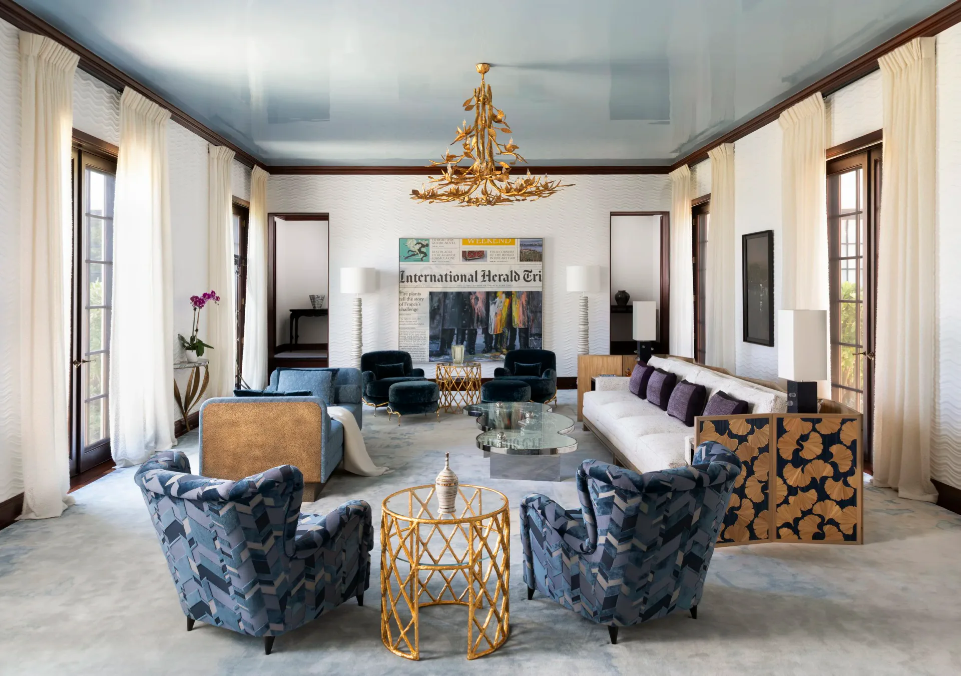 Luxurious living room with blue patterned armchairs, gold geometric side table, and fireplace, featuring cream curtains and modern artwork by Amanda Riha.