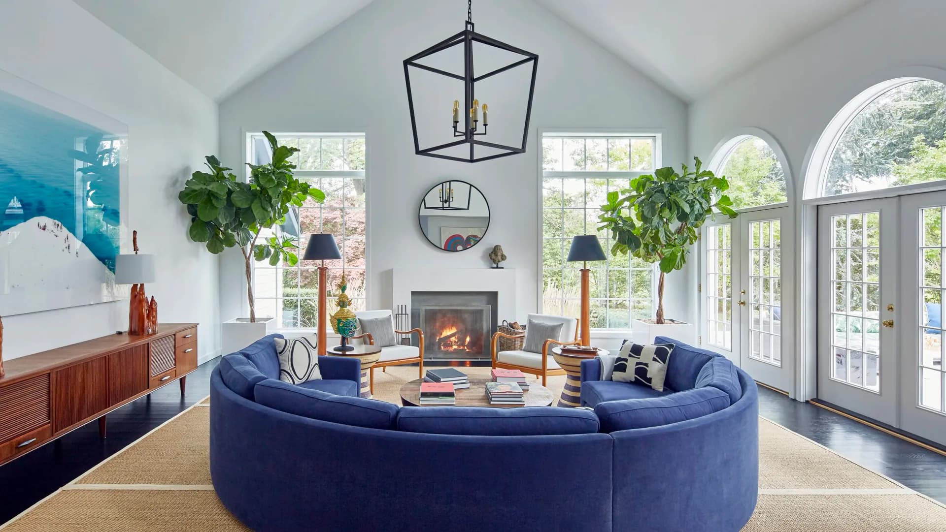 Modern living room with curved blue sectional sofa, fireplace, abundant natural light from arched windows, hanging plants, and contemporary furnishings by Amanda Riha.