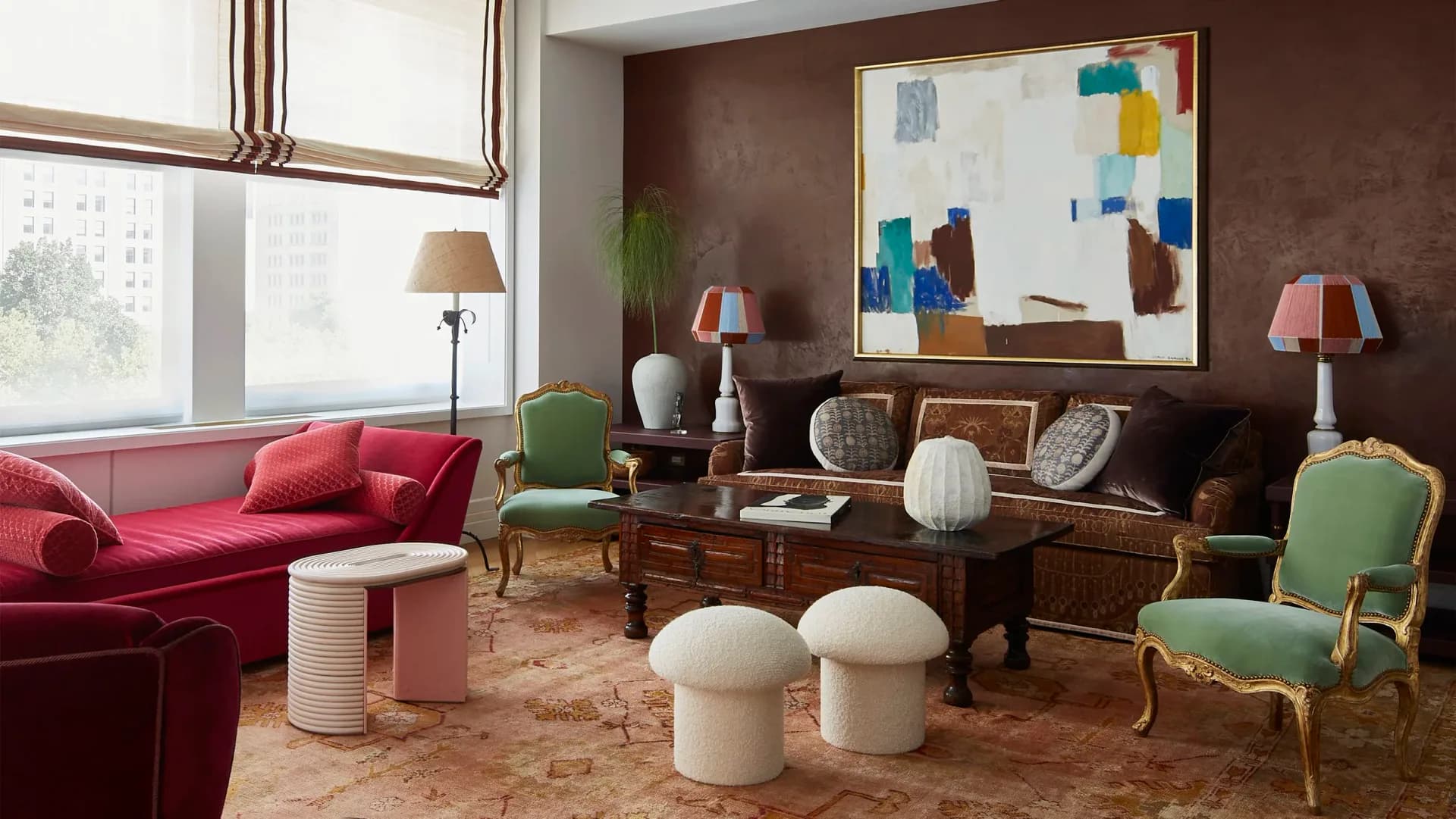 Luxe living room with chocolate brown walls, red velvet sofa, brass-framed chairs, patterned area rug, and modern abstract artwork by Amanda Riha.