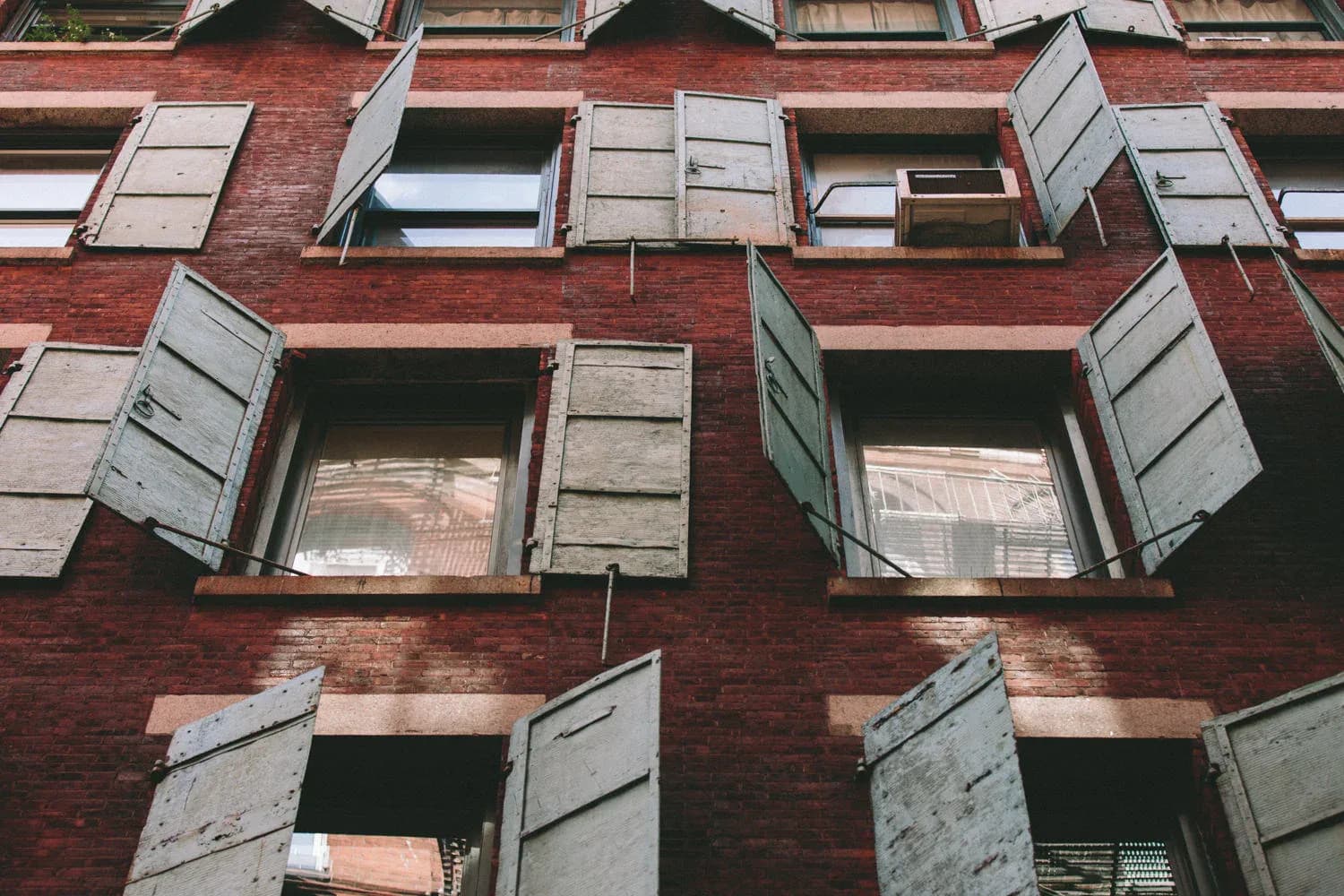 Red brick residential building facade with boarded-up windows and metal shutters, architectural documentation by Amber Troyer.