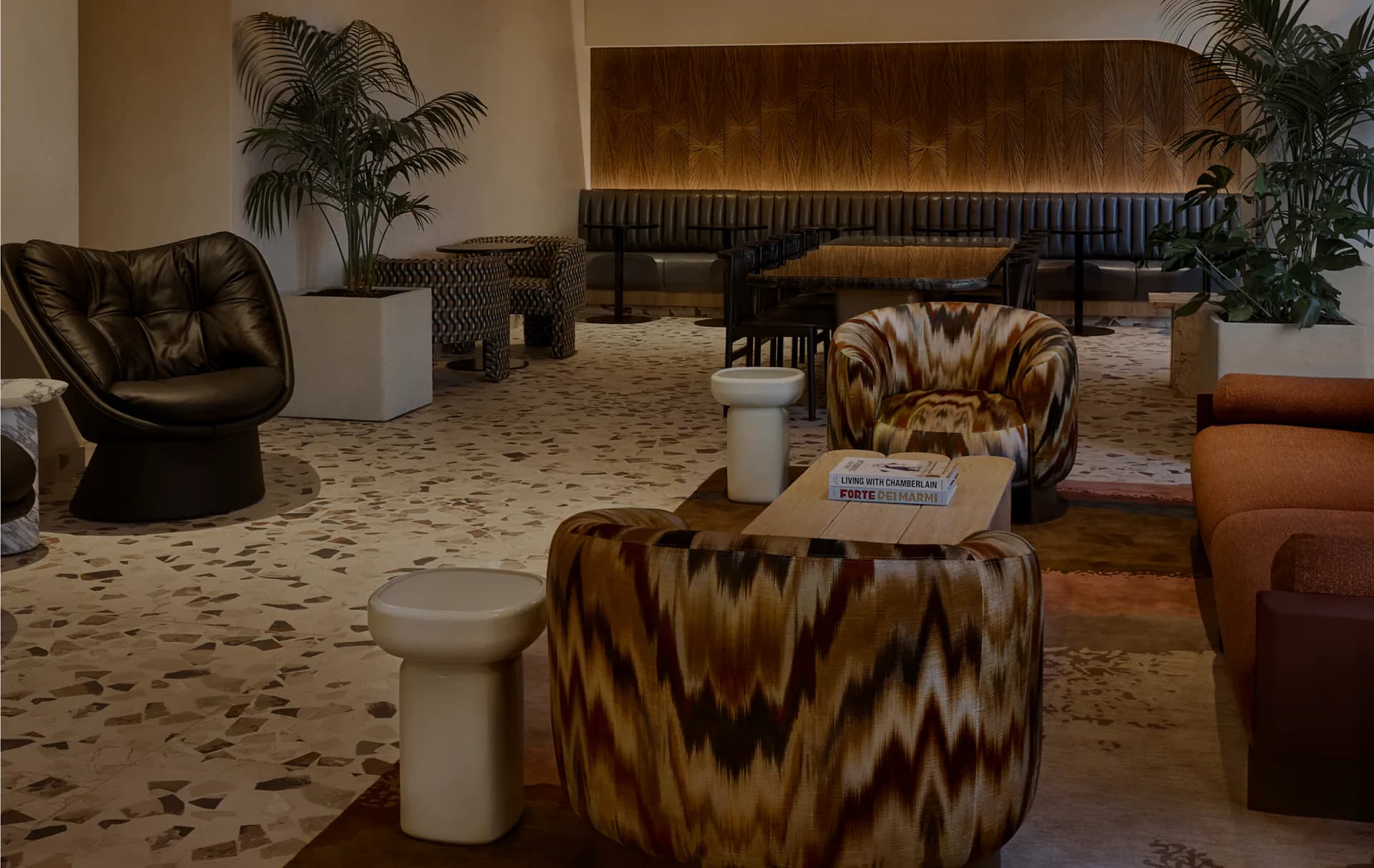 Warm-toned hotel lobby lounge with patterned accent chairs, curved seating, potted palms, and terrazzo flooring by Amelia Munning.