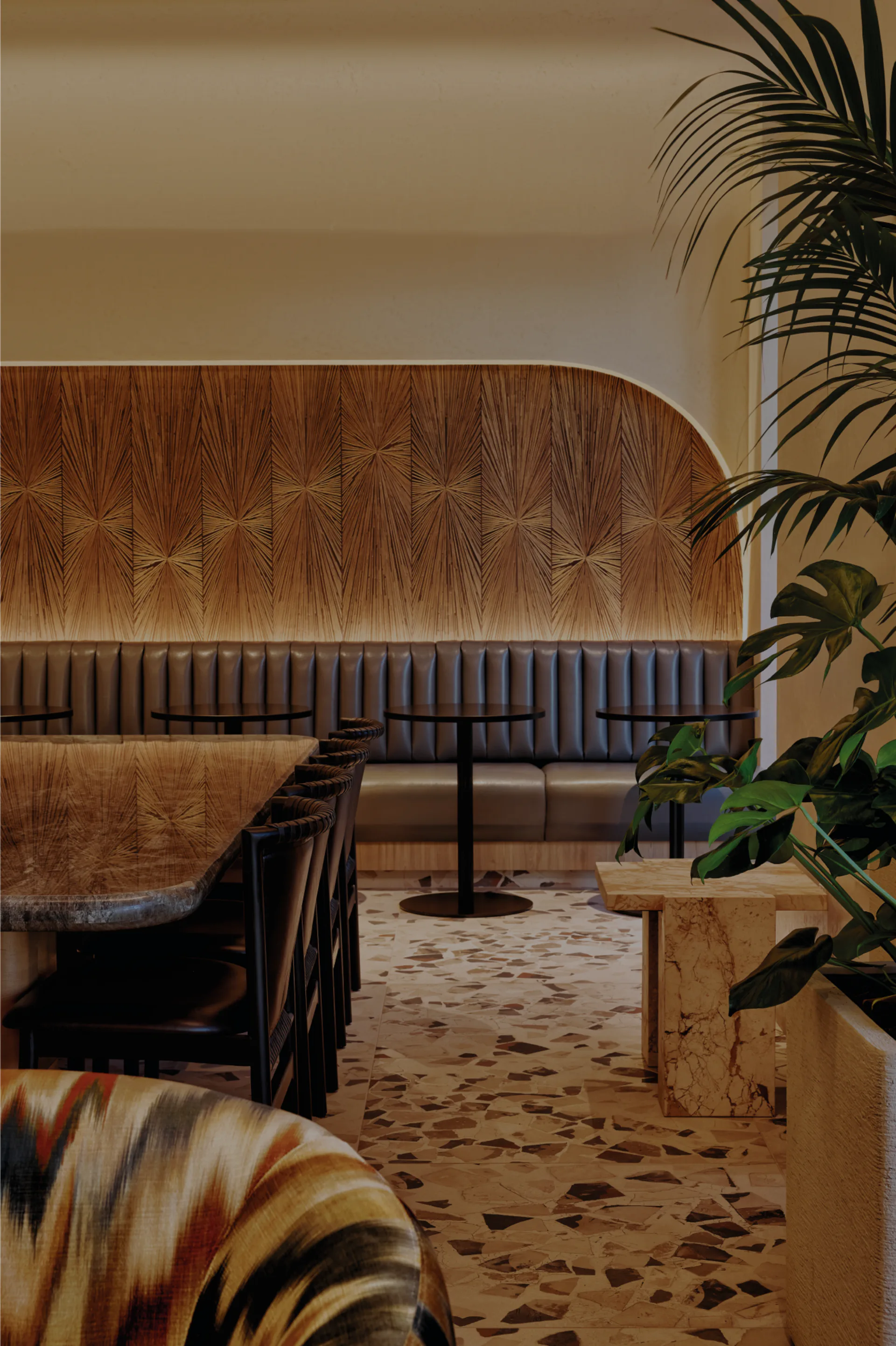 A warm-toned lounge interior featuring a curved wood-patterned banquette, striped blue seating, terrazzo flooring, and potted palms, designed by Amelia Munning.