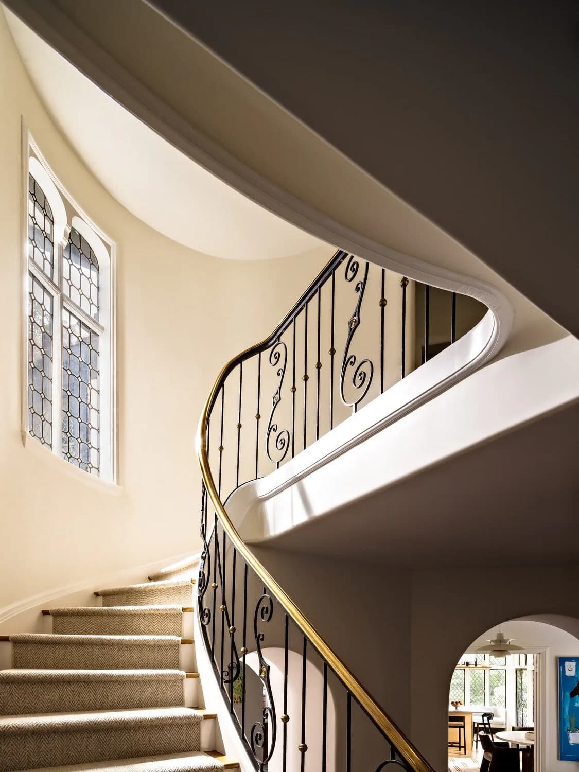 Curved spiral staircase with ornate iron railings and brass handrail, cream treads, and dramatic curved ceiling with skylight by Amy Courtney.