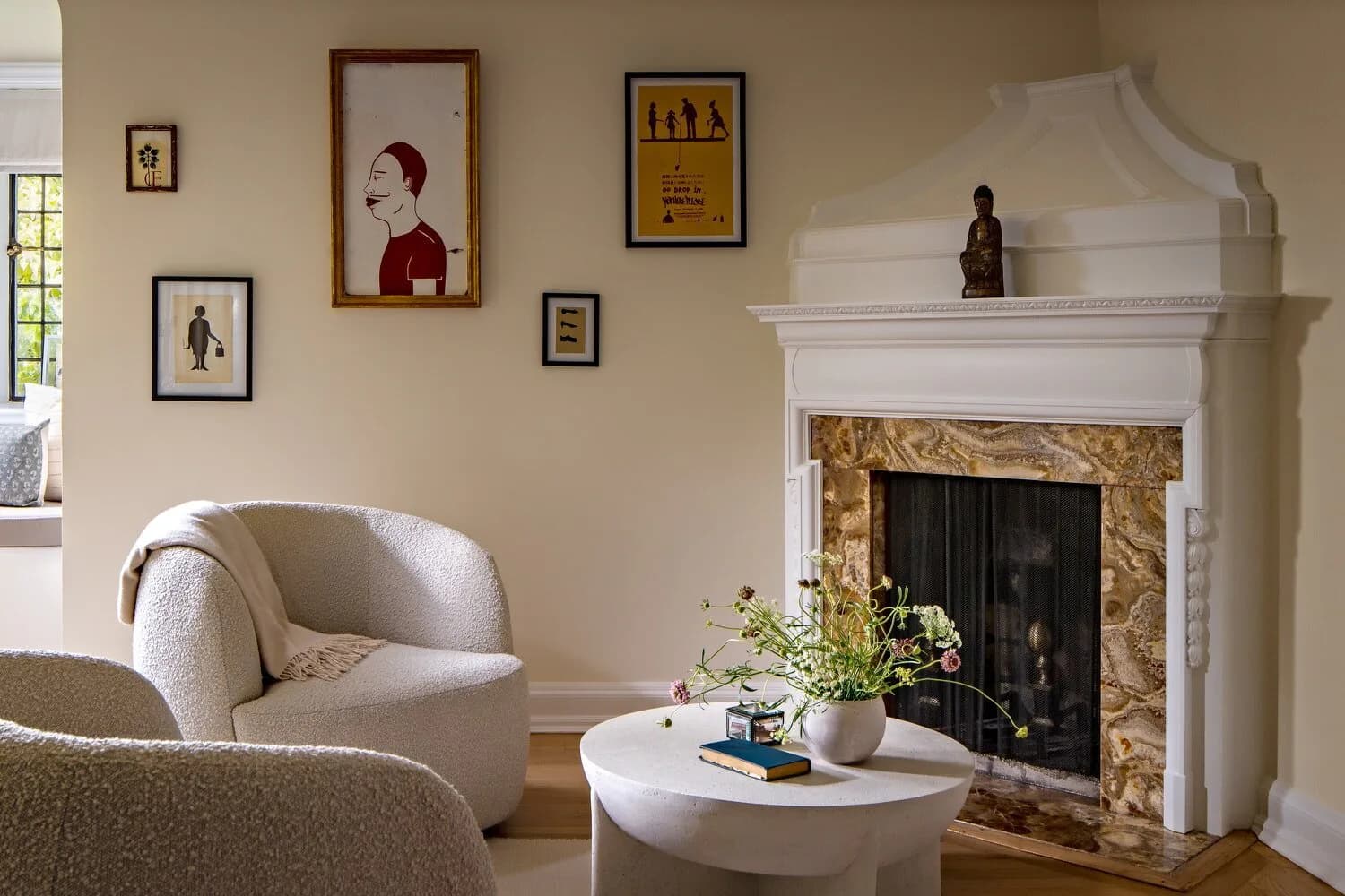 A serene living room with a curved cream armchair, round white coffee table, and ornate white fireplace with gold detailing, styled with framed artwork and greenery by Amy Courtney.
