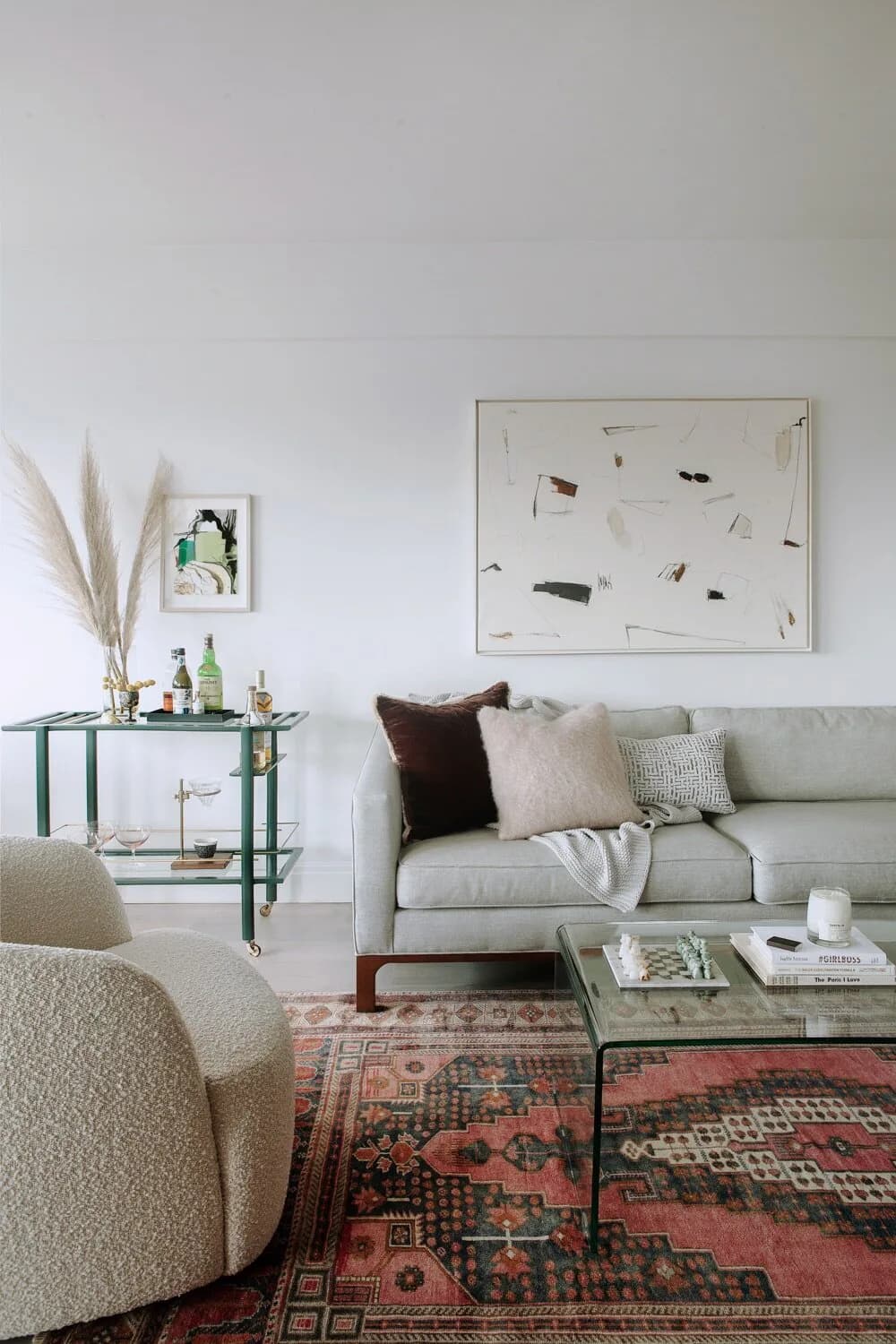 A minimalist living room features a gray sofa with layered pillows, a curved cream chair, patterned Turkish rug, green metal side table, and abstract wall art by Amy Courtney.