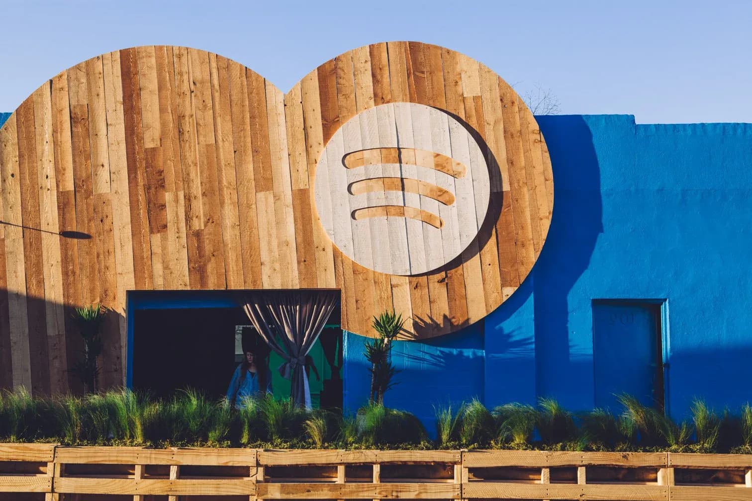 Wooden curved architectural installation with geometric cutout details and blue structural elements, against clear sky by Amy Gavin.