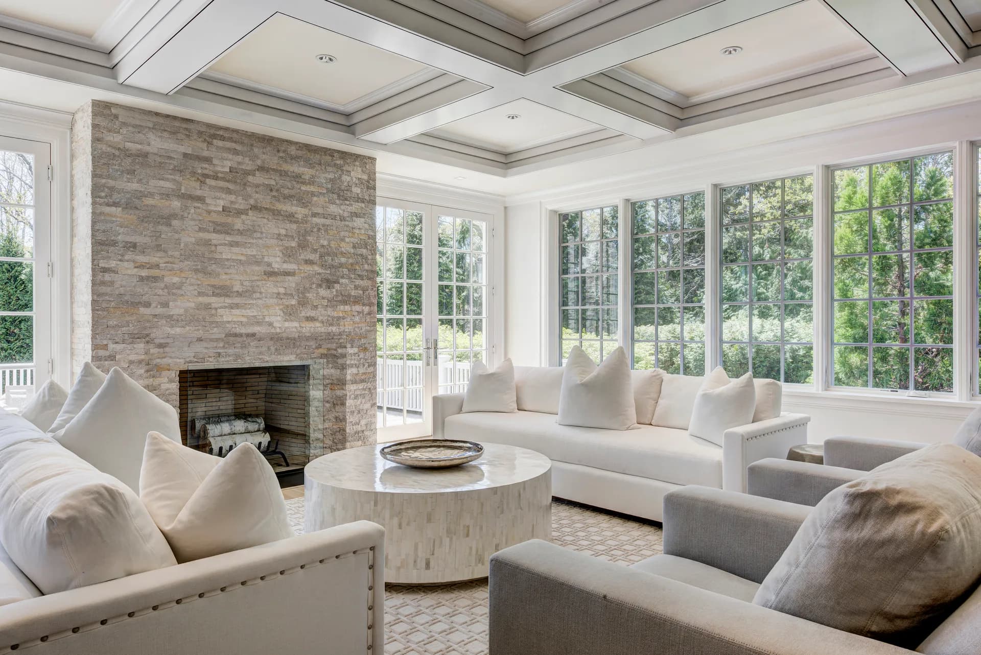 Bright luxury living room with coffered ceiling, brick fireplace, white furnishings, round marble table, and expansive windows overlooking greenery by Amy Hill.