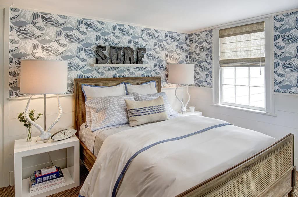 Coastal-inspired bedroom with blue wave-patterned wallpaper, wooden bed frame, white nightstands, and striped bedding in blue and white tones by Amy Hill.