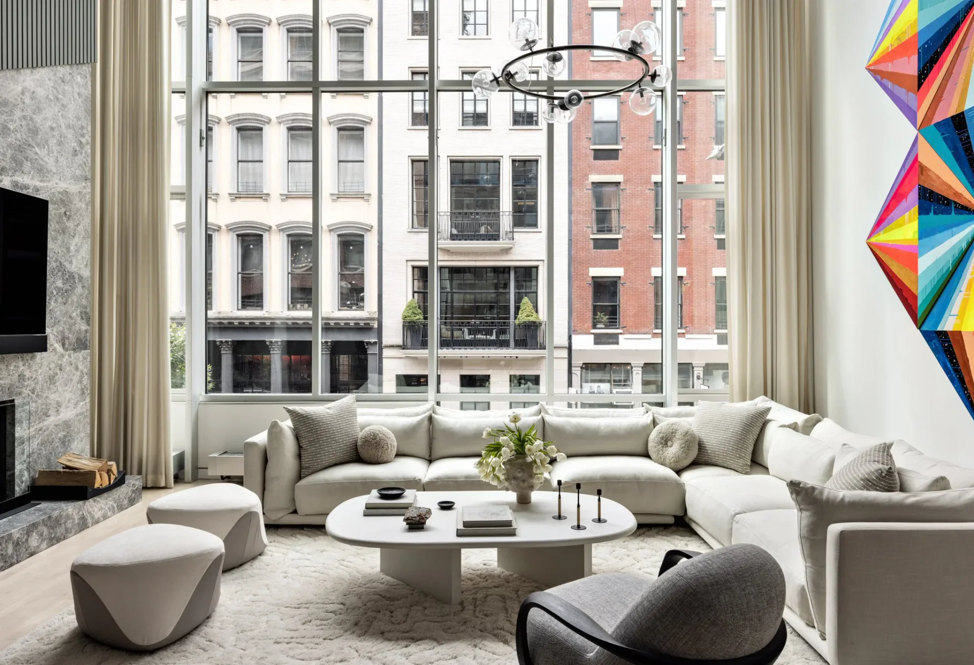 A modern living room with curved sectional sofa, round marble table, and floor-to-ceiling windows overlooking NYC buildings, designed by Amy Kalikow.
