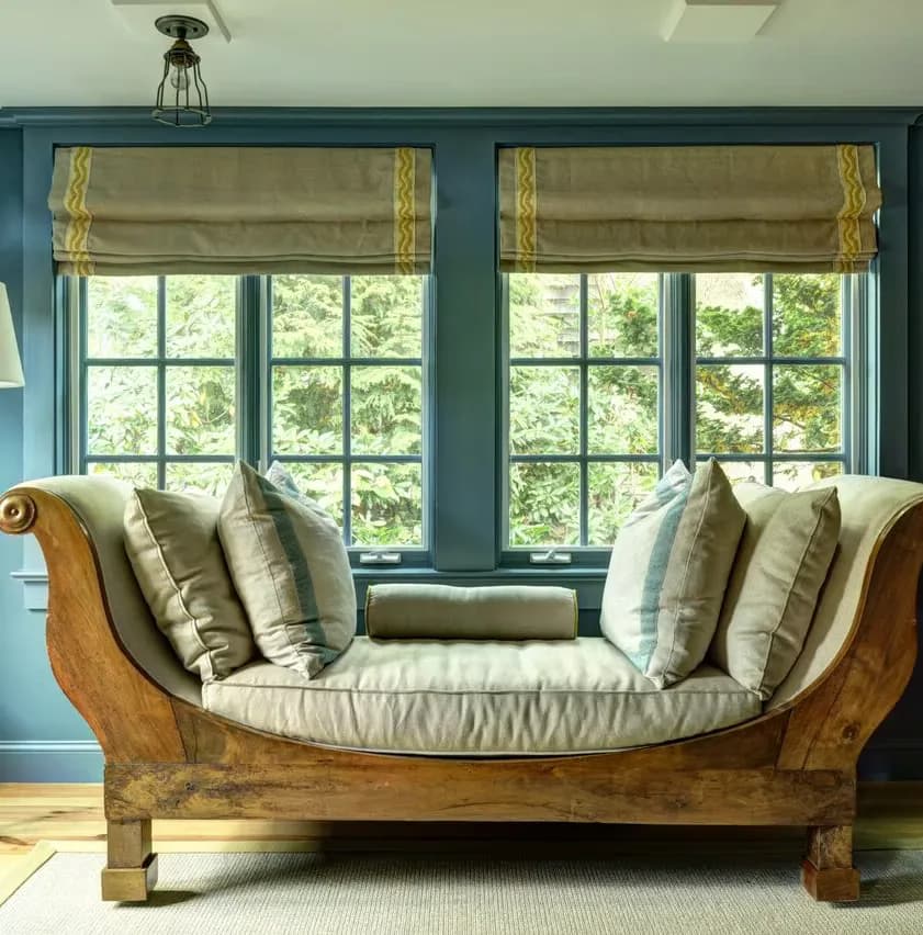 A teal-walled window seat features a wooden bench with cream cushions and patterned pillows, framed by gold-trimmed roman shades overlooking lush greenery, designed by Amy Werfel.