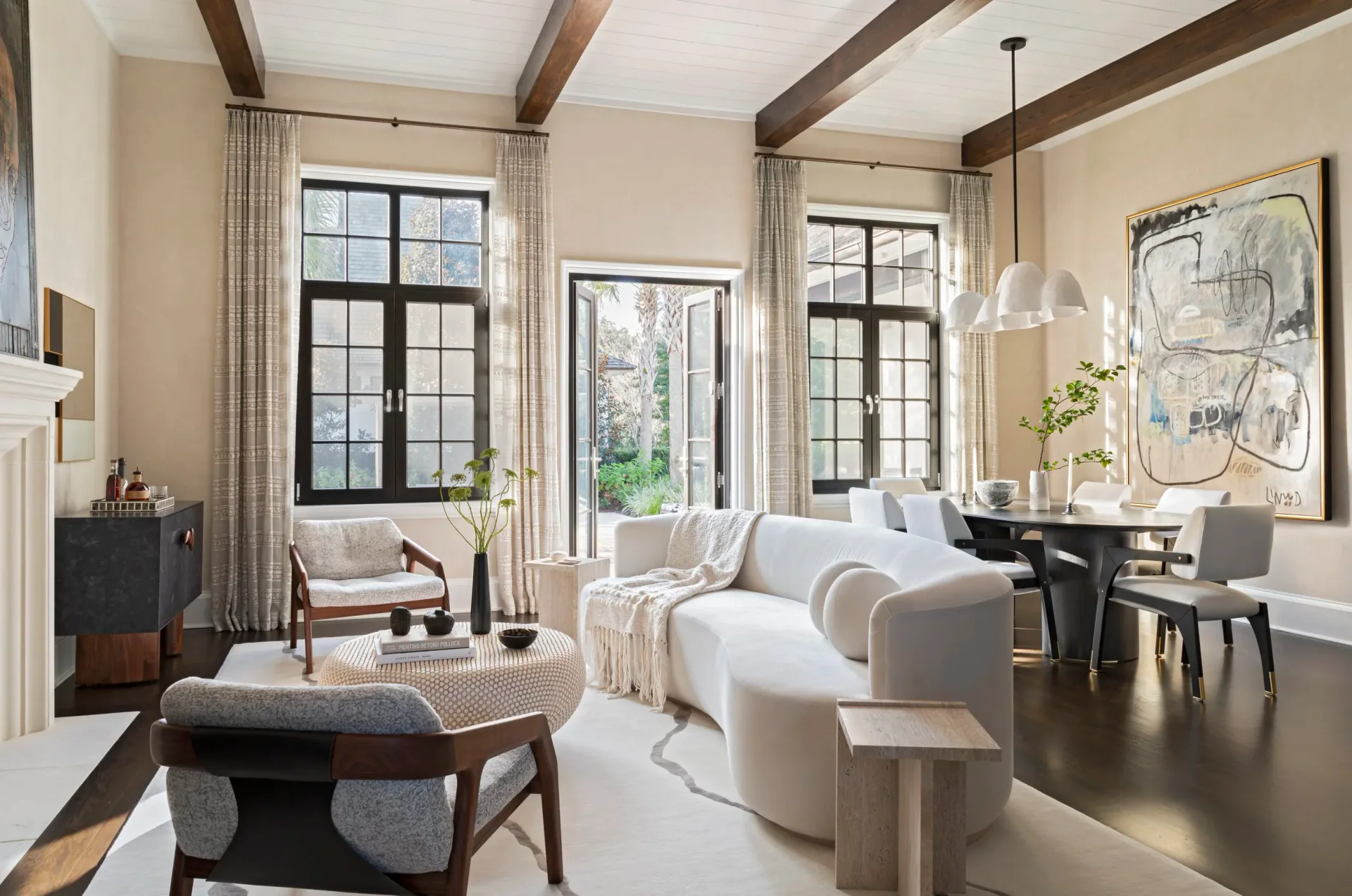 Luminous living room with curved cream seating, dark wood beams, multiple windows with curtains, and modern furnishings in warm neutrals by Ana Junco.