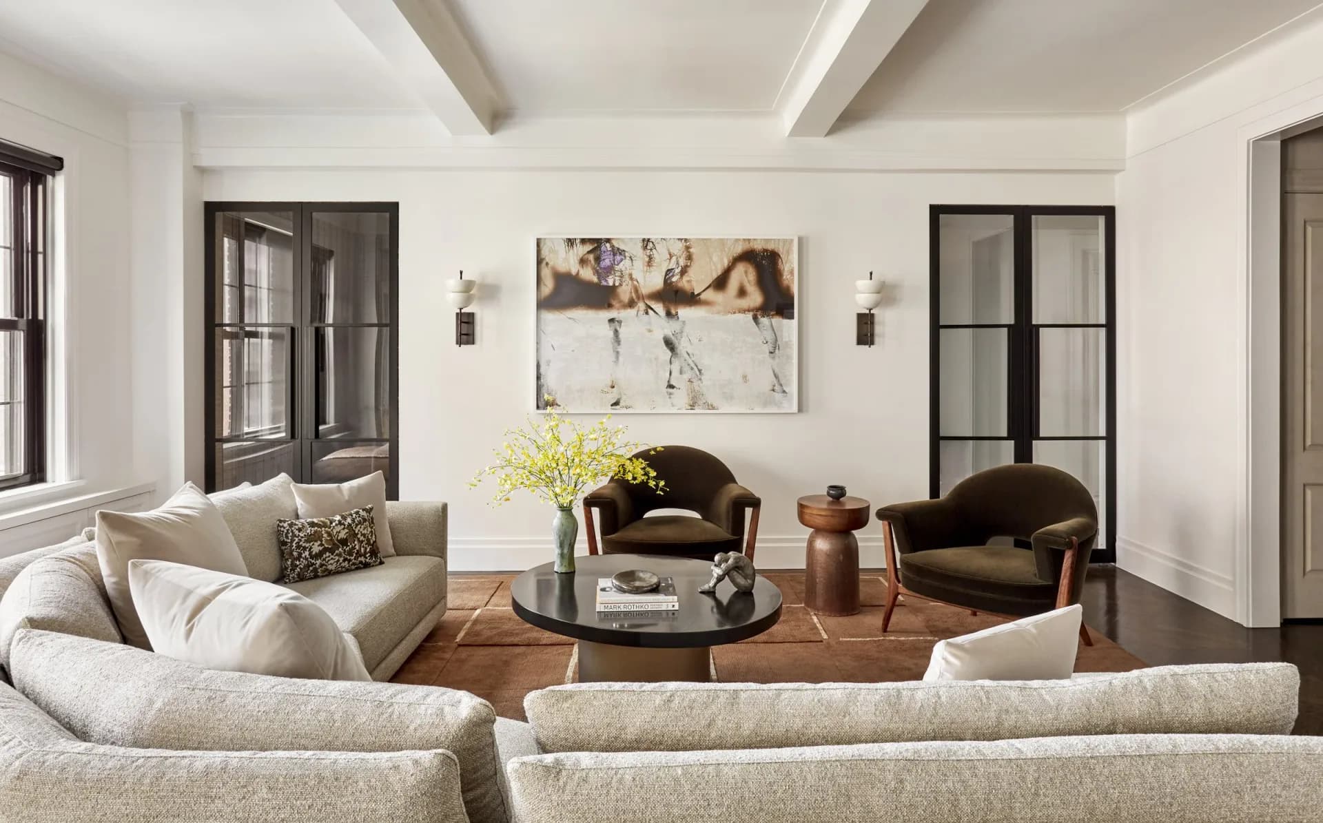 Sophisticated living room with cream curved sectional, dark accent chairs, black round coffee table, and exposed beam ceiling by Ana Junco.