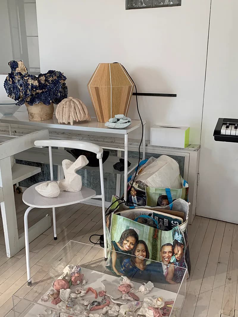 Styled interior vignette featuring a round white side table with white sculptural objects, wooden coffin-shaped decorative piece, and potted plant arrangement on upper shelving by Ana Kraš.