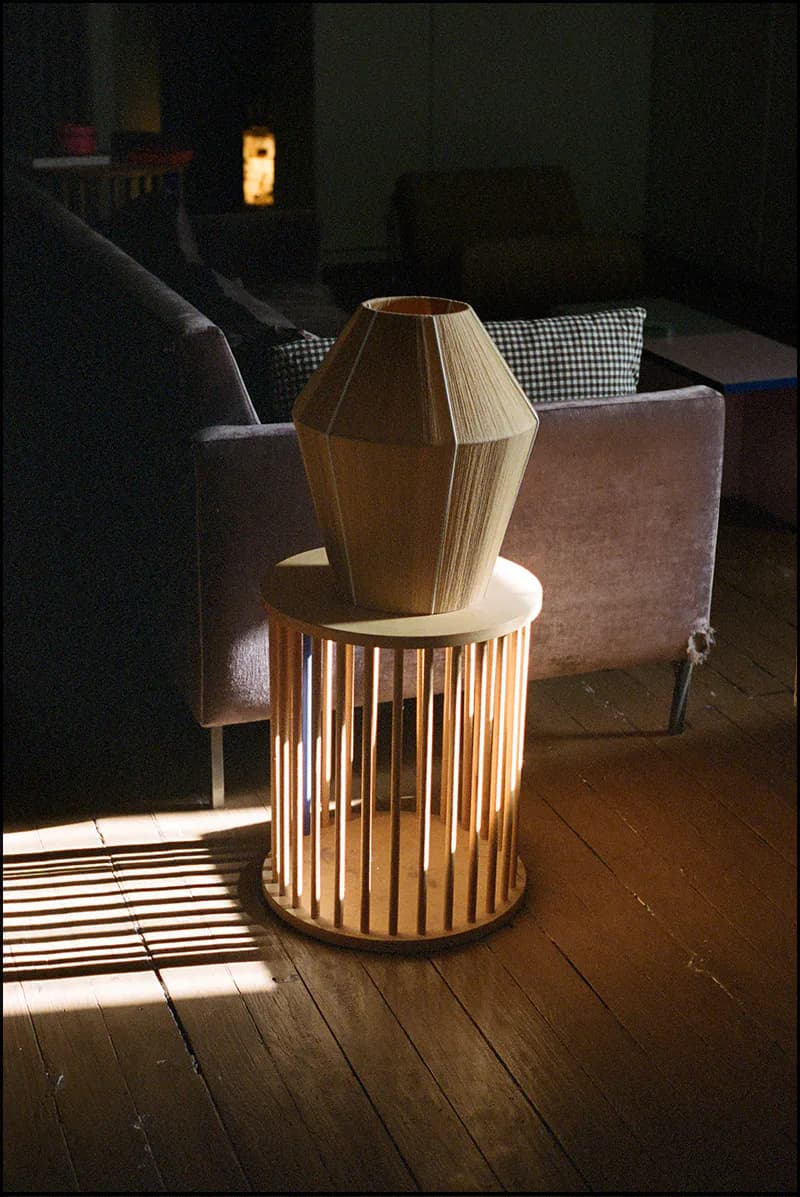 A wooden geometric pendant light suspended above a cylindrical slatted wooden base, illuminated by warm sunlight in a gallery space by Ana Kraš.