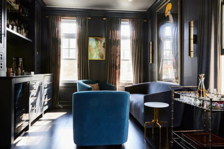 Luxurious dark lounge with deep blue curved armchair, black walls, brass accents, and tall windows with dramatic drapery, designed by Ana-Maria Leeming.