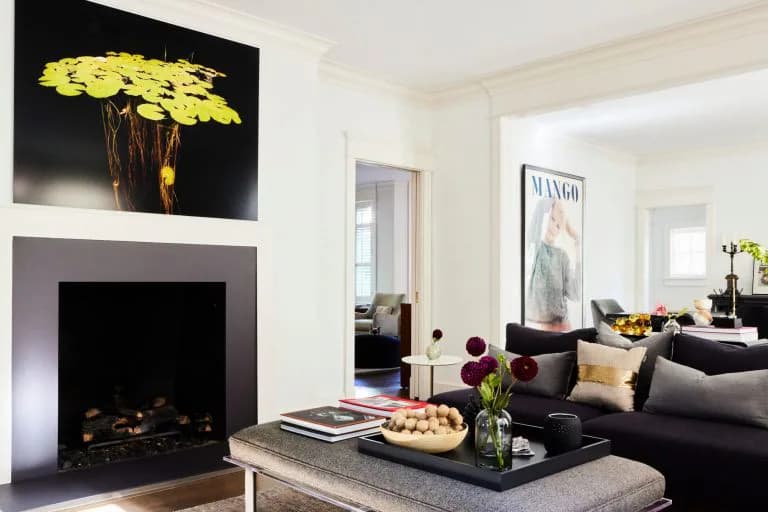 Modern living room with black fireplace surround, gray concrete hearth, black sofa, yellow floral artwork, and minimalist furnishings by Ana-Maria Leeming.