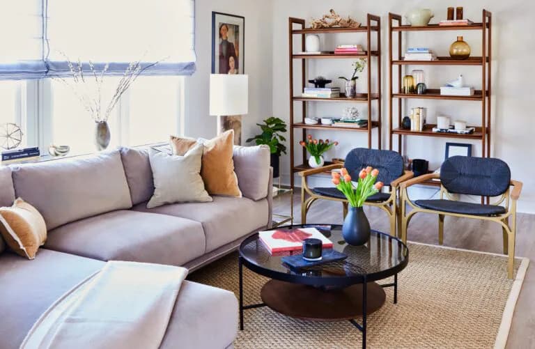 Modern living room with gray sectional sofa, black round coffee table, wooden shelving units, and blue accent chairs, designed by Ana-Maria Leeming.