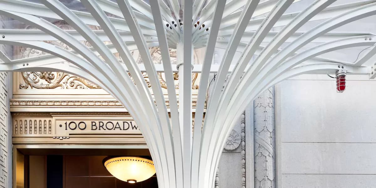 Elegant modern lobby featuring dramatic white geometric ceiling coffers, brass signage reading '100 BROAD', and refined architectural details by Ana Rojas.