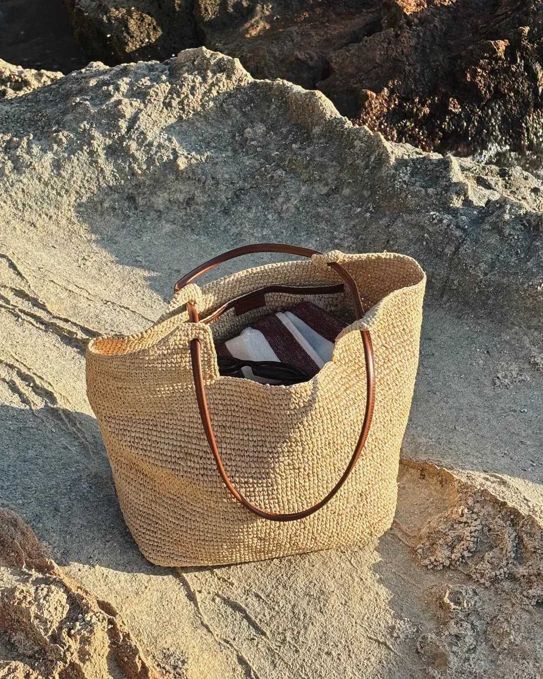 Natural woven straw tote bag with leather handles displayed on rocky coastal terrain by Analise Buhr.