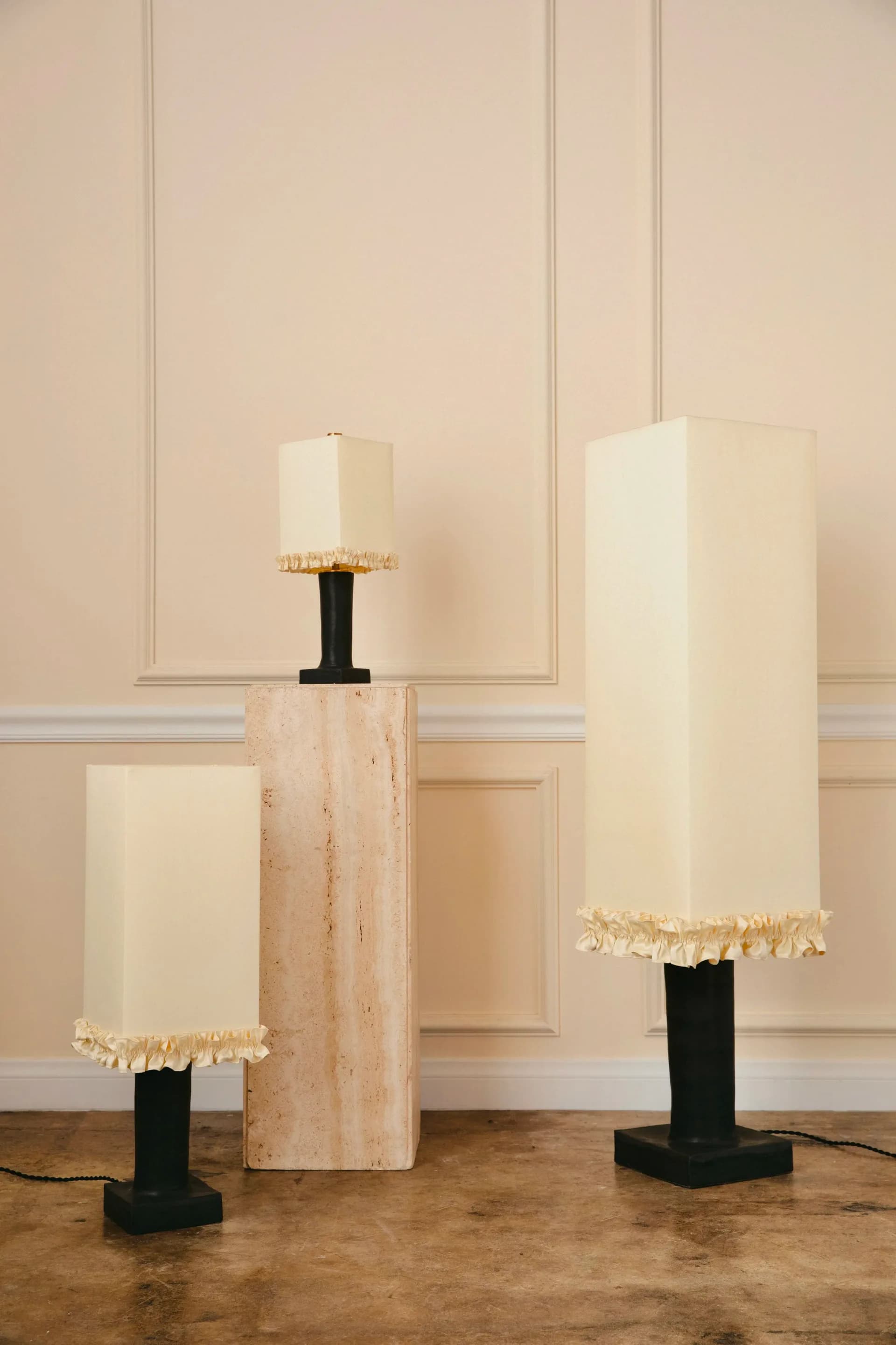 Three cylindrical table lamps with cream linen shades and gold-trimmed bases on black pedestals and travertine base, displayed against paneled beige wall by Analuisa Corrigan.